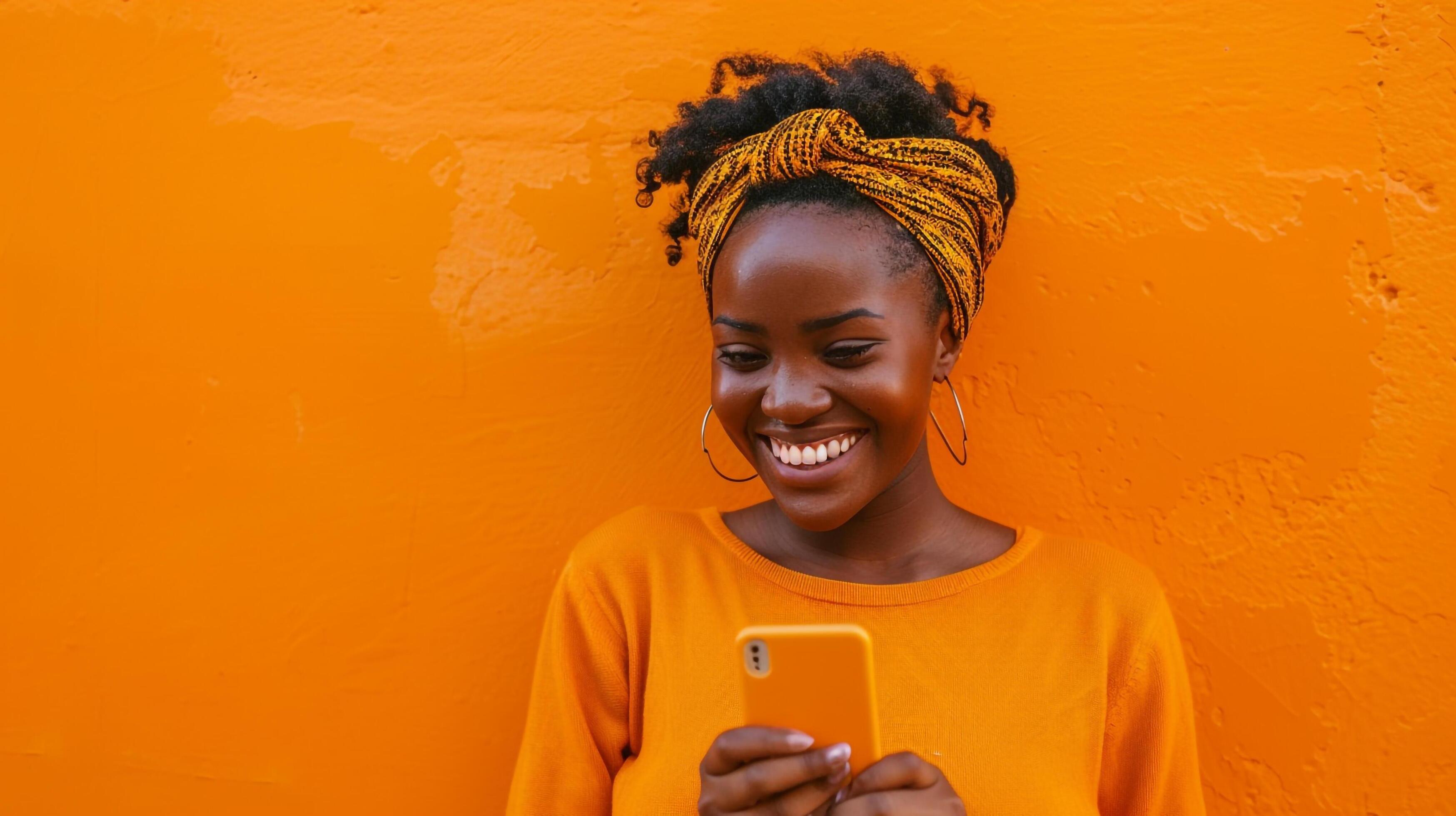 Happy lady in orange top texting by orange wall Stock Free