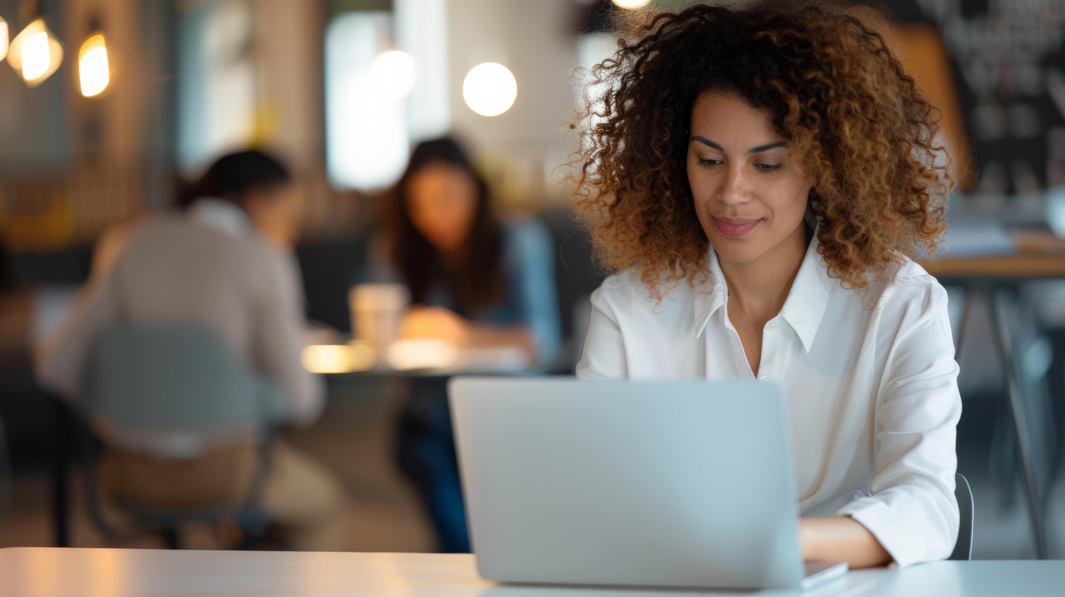 Young professional woman working on laptop in modern office setting with colleagues in the background, focused and productive Stock Free