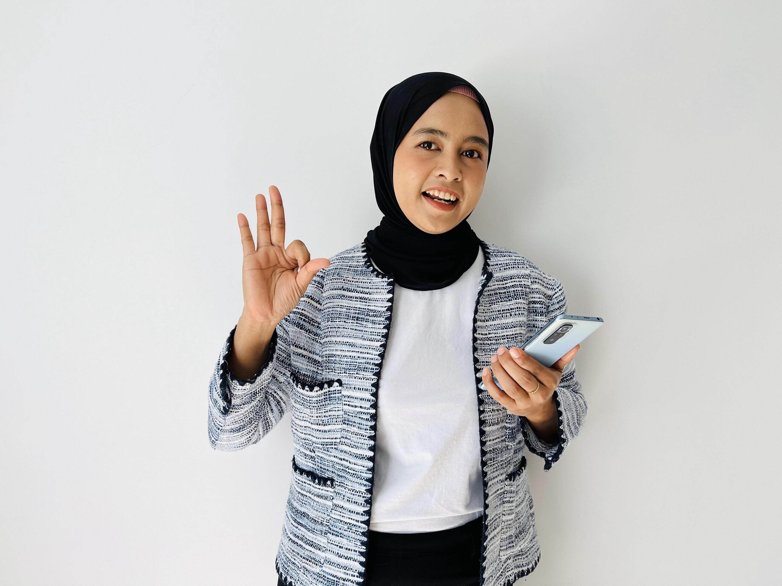 Portrait of smiling Asian Muslim woman OK gesture with the smartphone wearing black hijab and tweed blazer over white background Stock Free