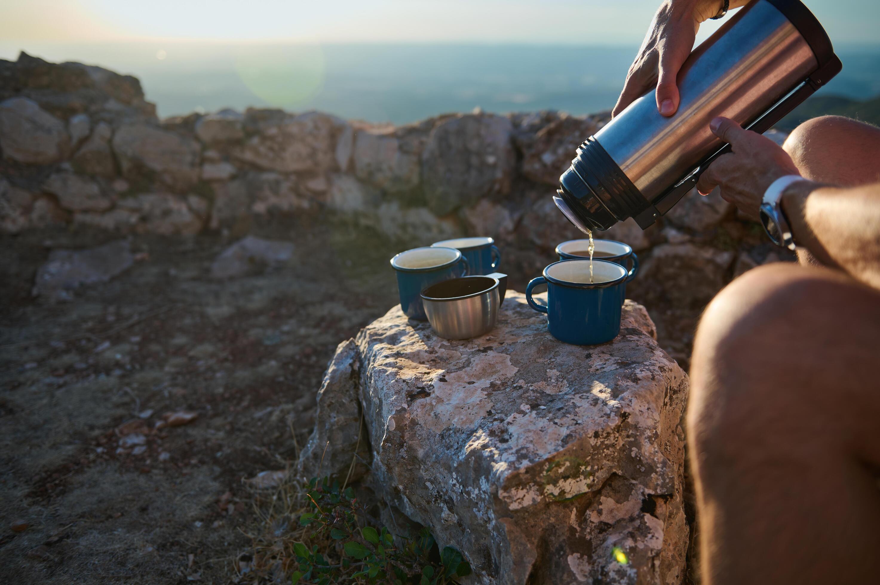 Person pouring coffee from thermos into cups during outdoor adventure, with rocky landscape in background Stock Free