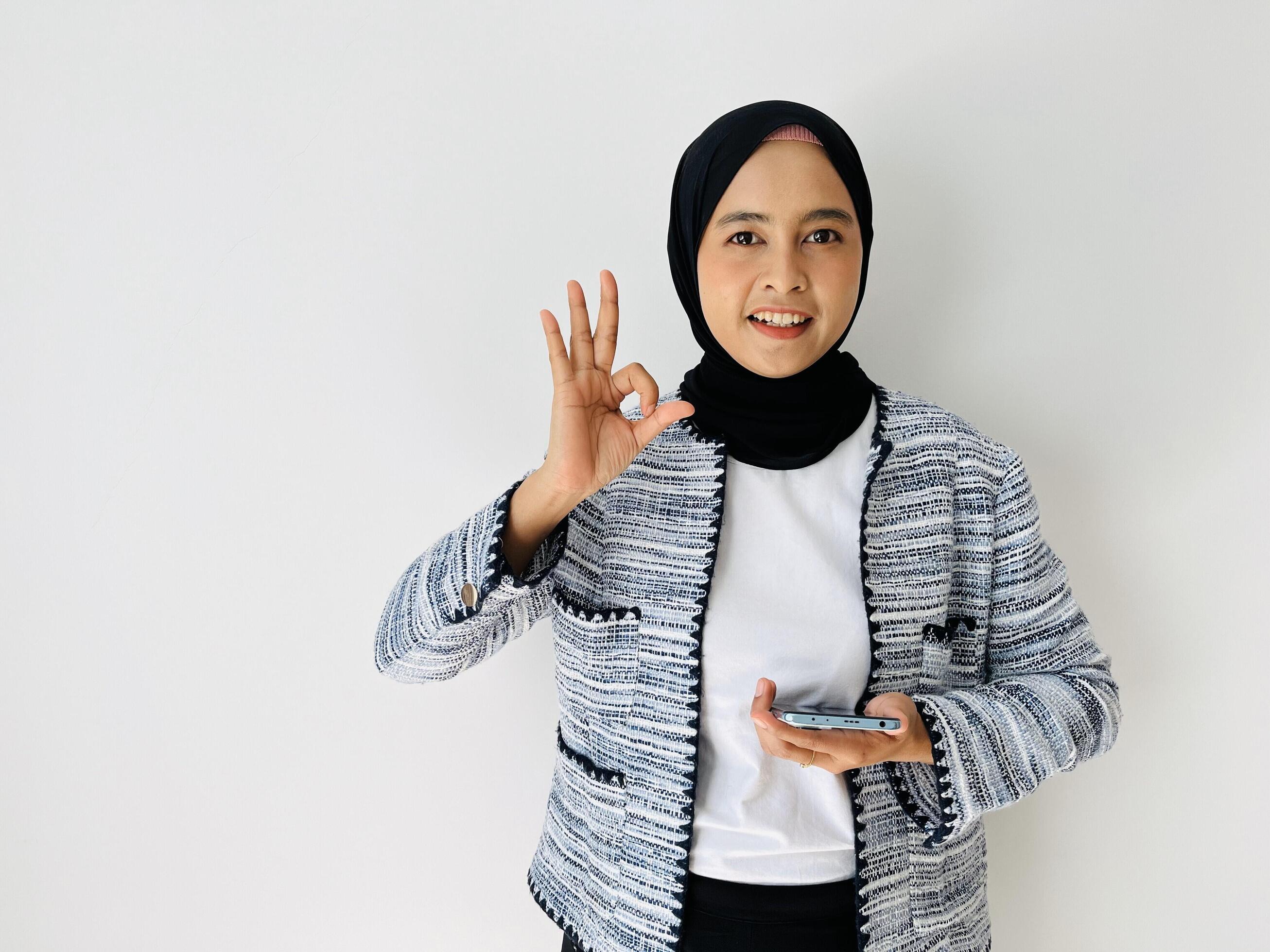 Portrait of smiling Asian Muslim woman OK gesture with the smartphone wearing black hijab and tweed blazer over white background Stock Free