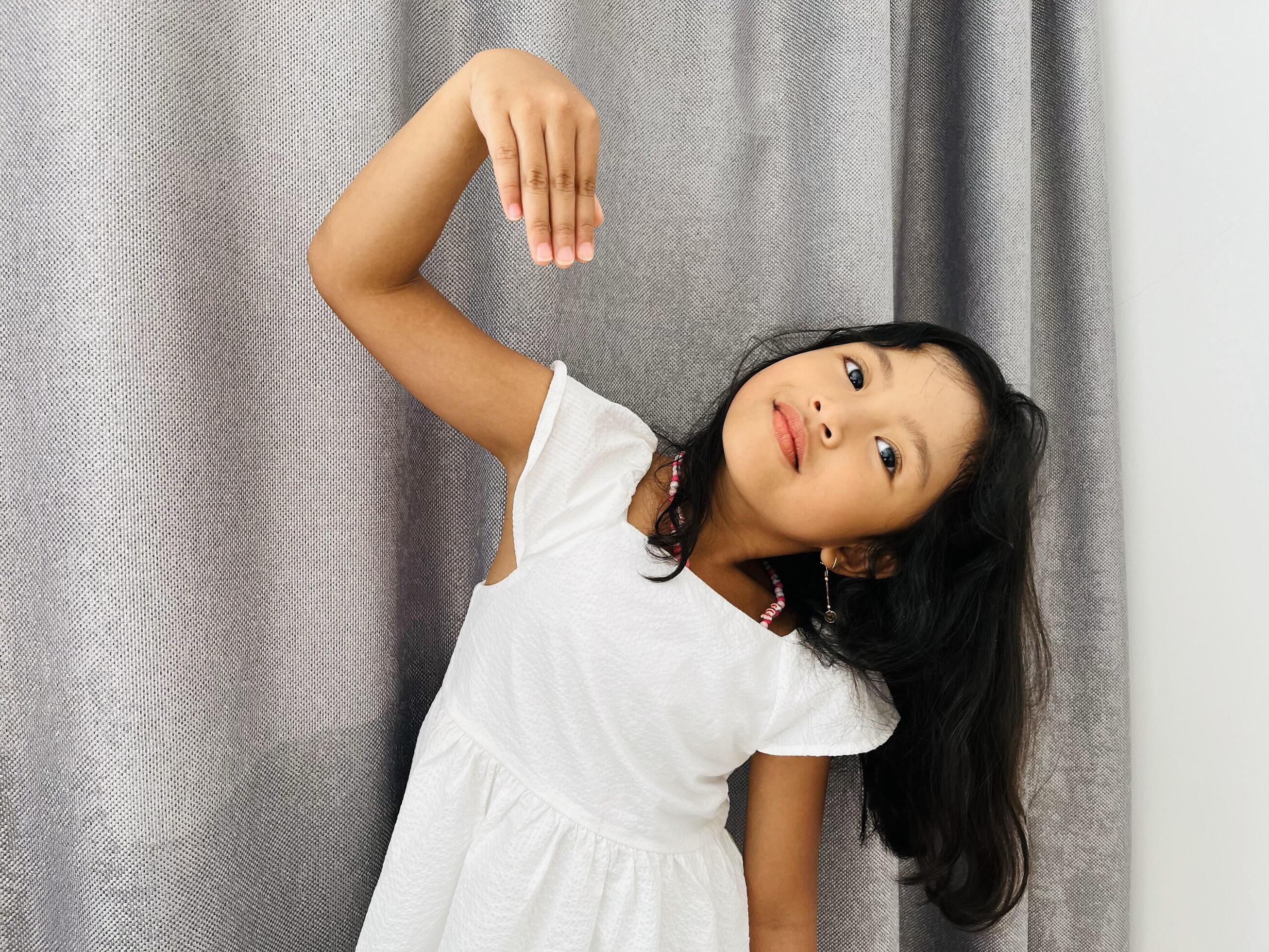 A five years old Asian child smiling look at the camera and one hand make half love sign near the grey curtain Stock Free