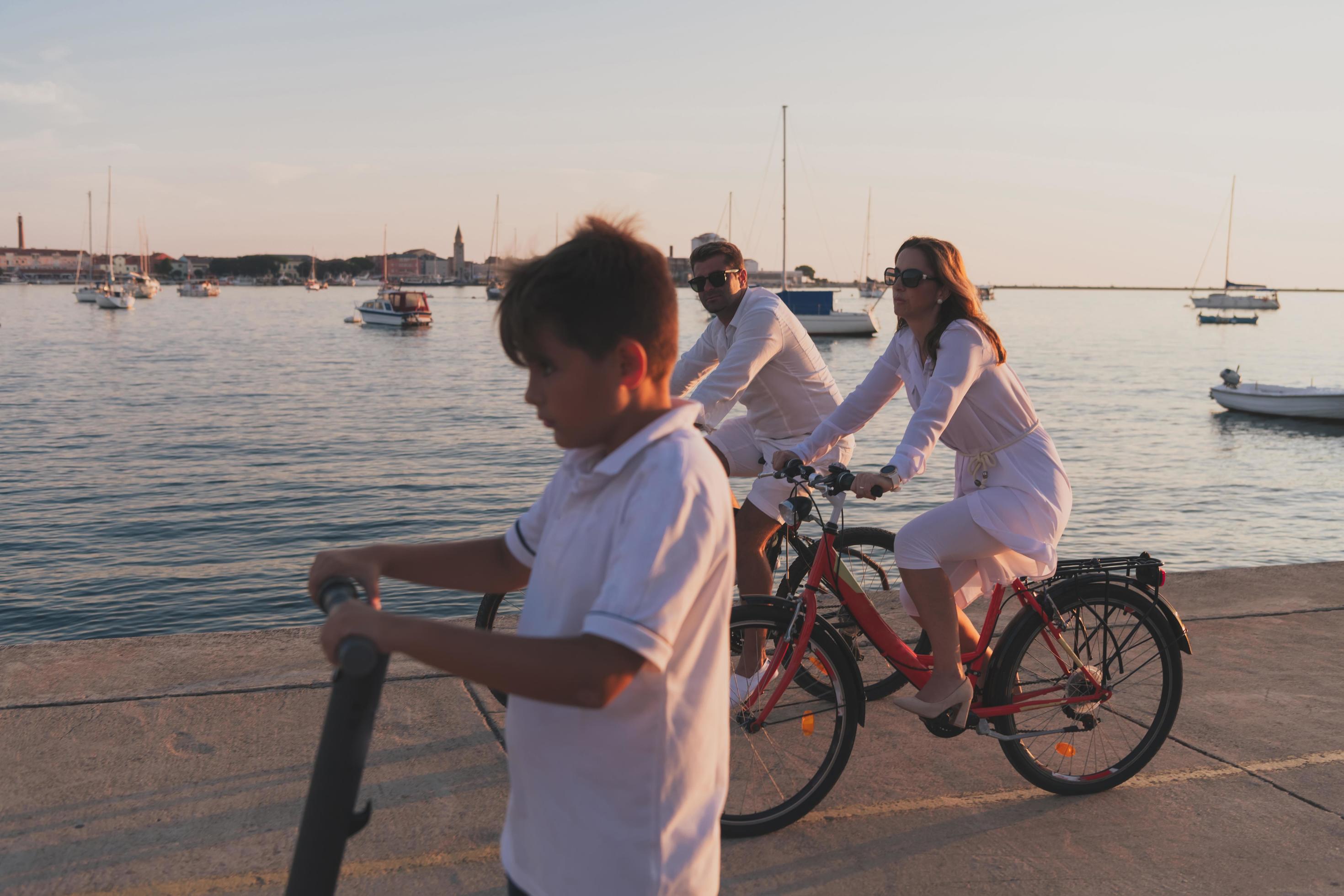 Happy family enjoying a beautiful morning by the sea together, parents riding a bike and their son riding an electric scooter. Selective focus Stock Free