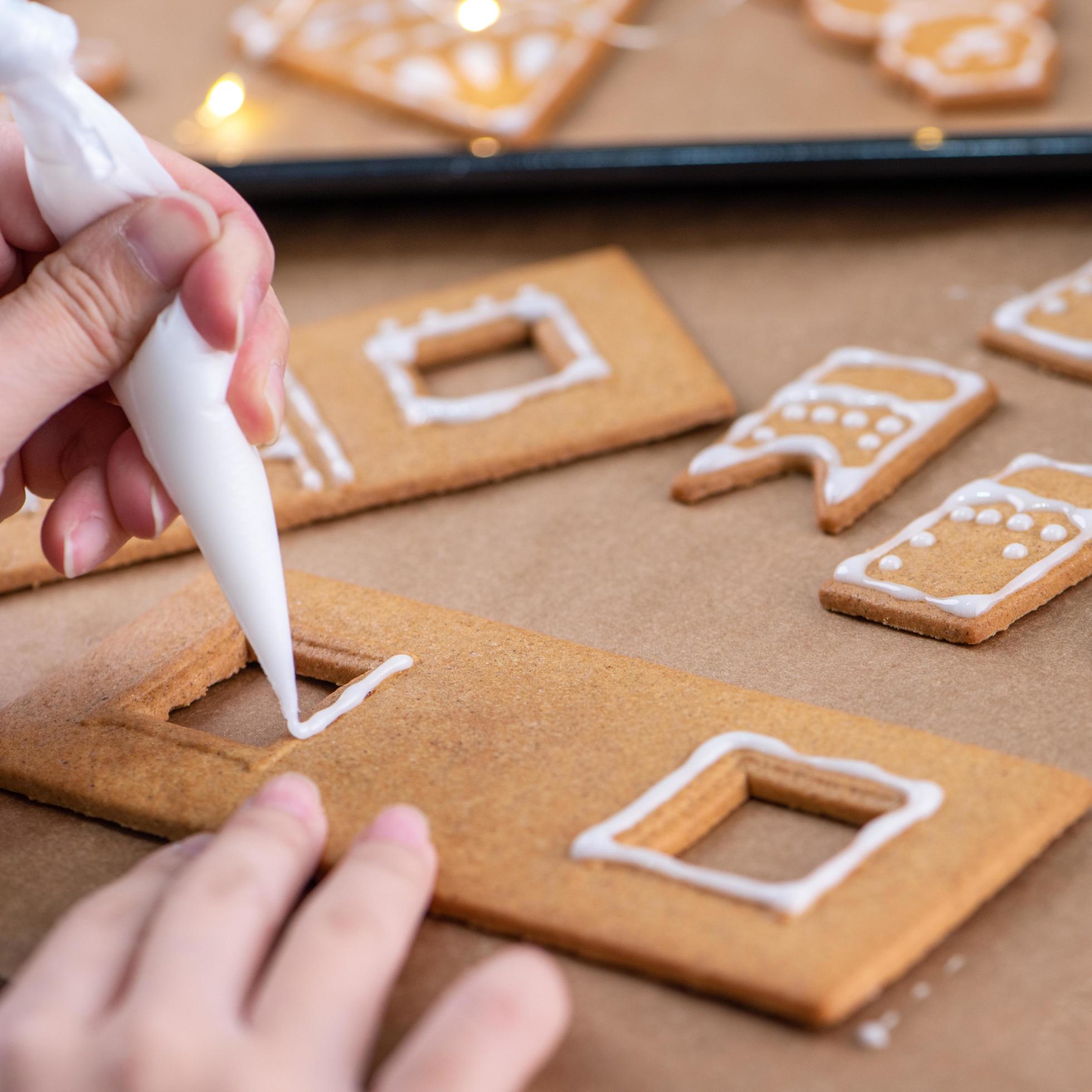 Young woman is decorating Christmas Gingerbread House cookies biscuit at home with frosting topping in icing bag, close up, lifestyle. Stock Free