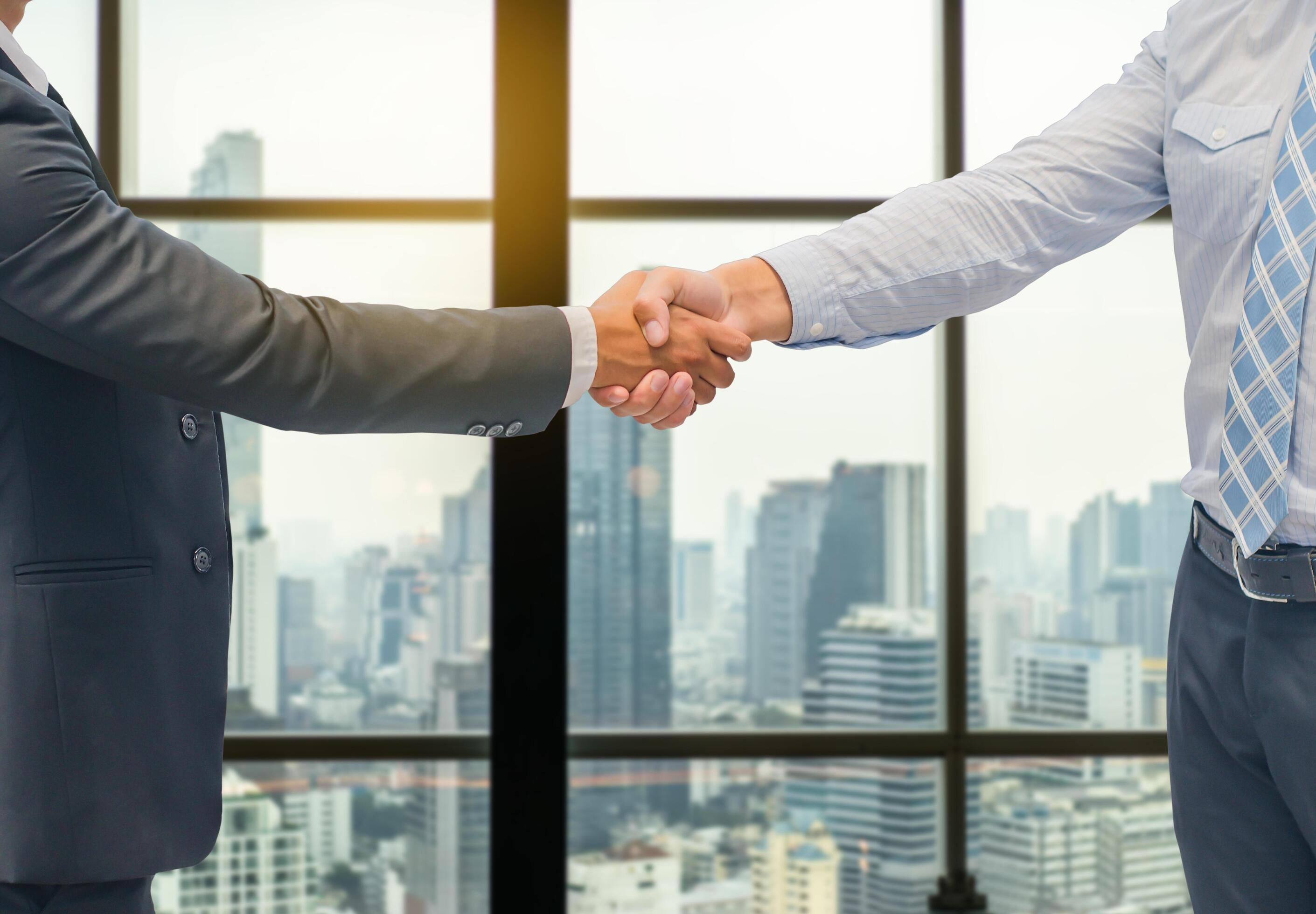 Close up of a business hand shake between two colleagues – greeting, dealing, merger and acquisition concepts on Modern glass building background Stock Free