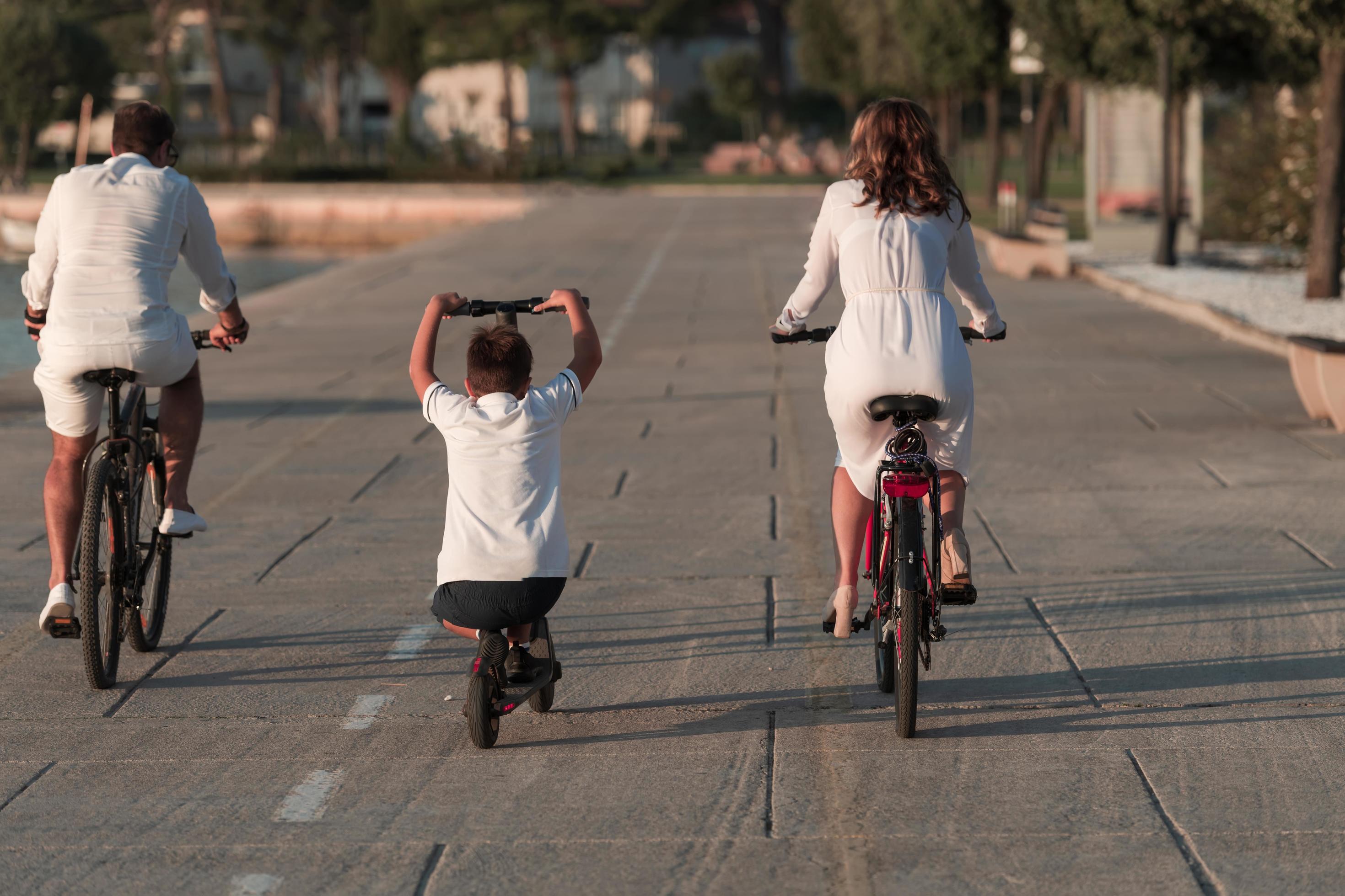 Happy family enjoying a beautiful morning by the sea together, parents riding a bike and their son riding an electric scooter. Selective focus Stock Free