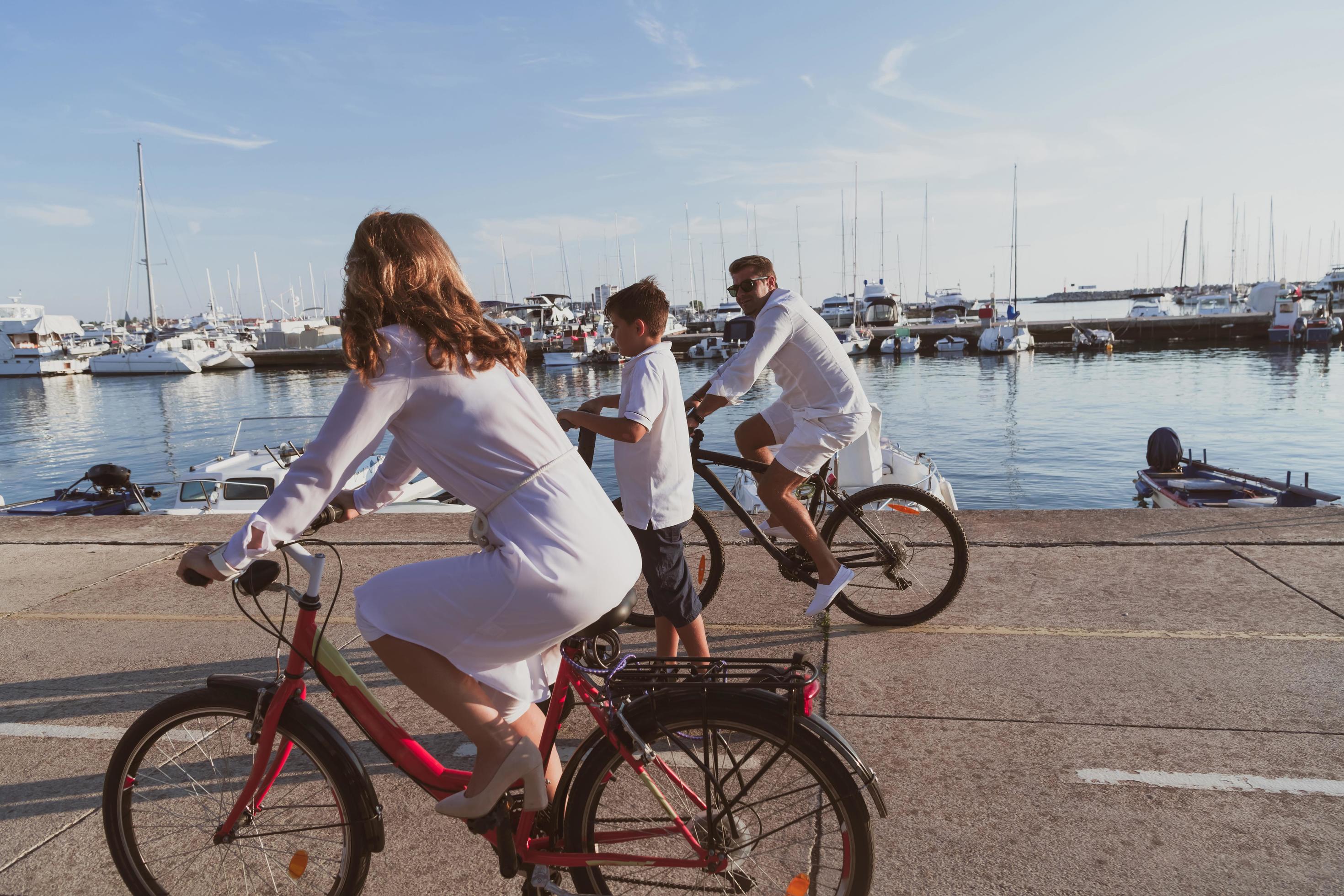 Happy family enjoying a beautiful morning by the sea together, parents riding a bike and their son riding an electric scooter. Selective focus Stock Free