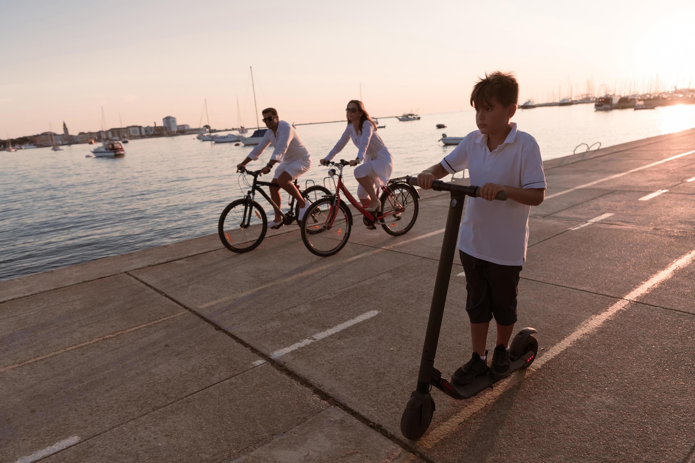 Happy family enjoying a beautiful morning by the sea together, parents riding a bike and their son riding an electric scooter. Selective focus Stock Free