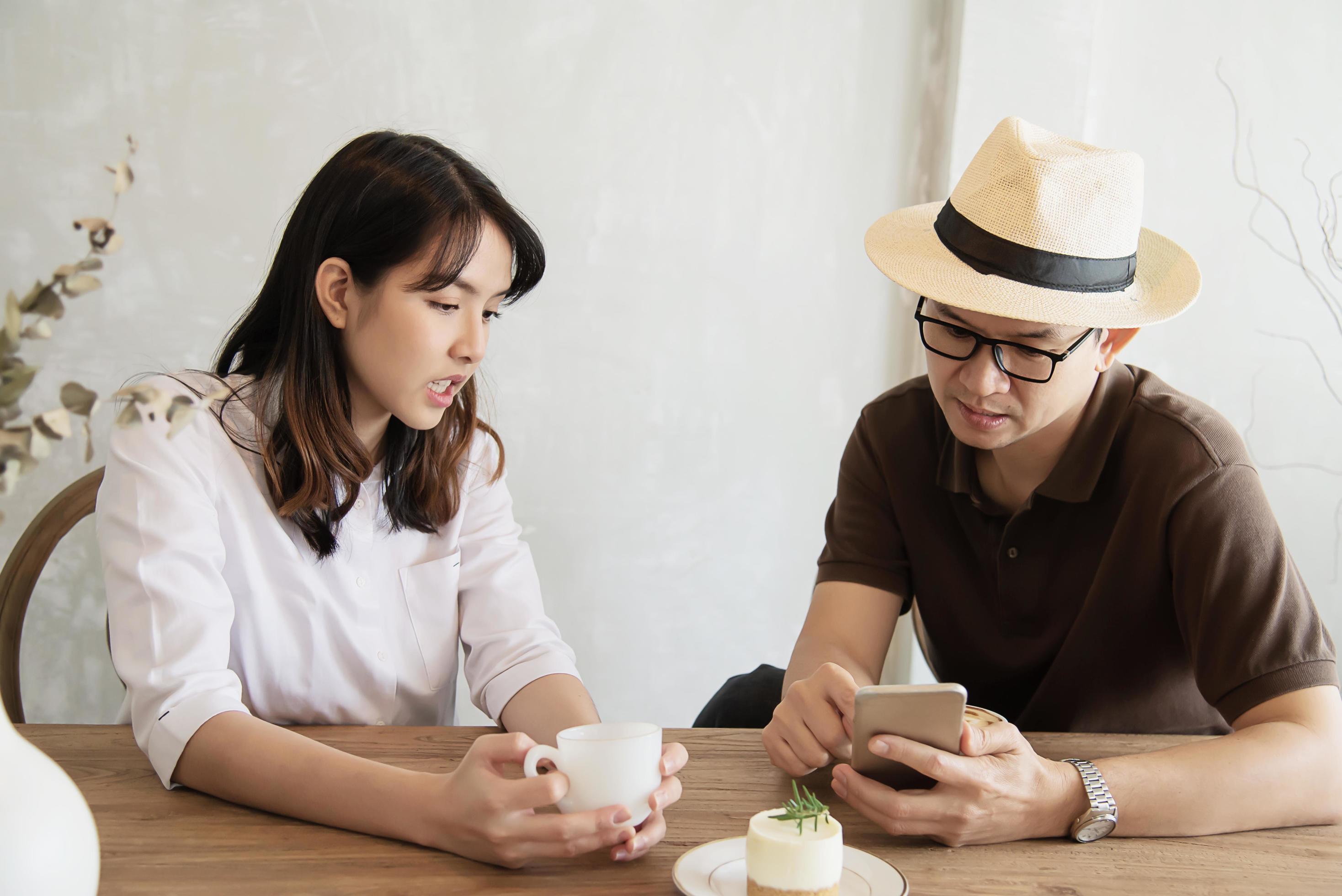 Casual man and woman talking happily while drink coffee and looking mobile phone – happy time lifestyle in coffee shop Stock Free