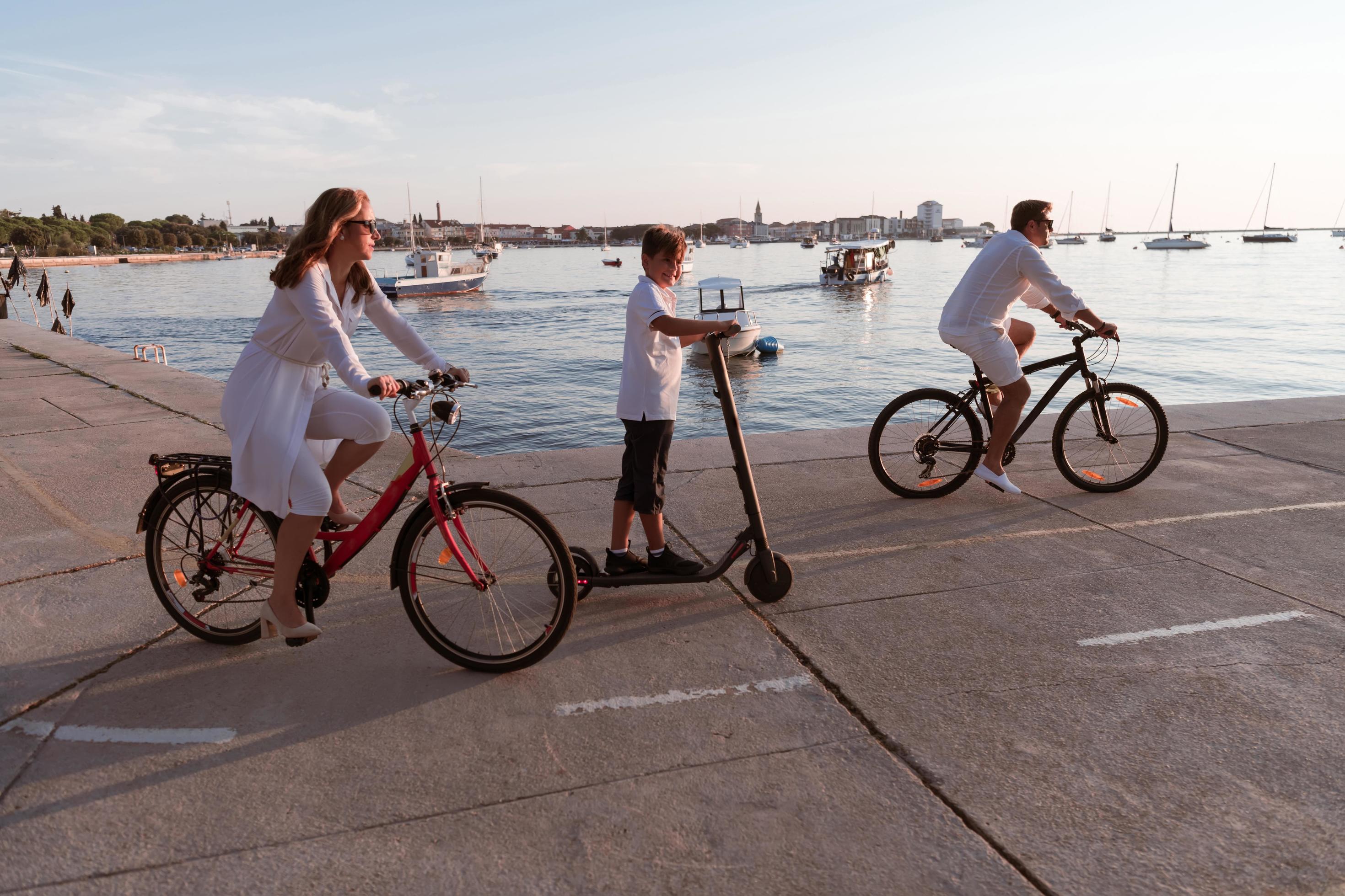 Happy family enjoying a beautiful morning by the sea together, parents riding a bike and their son riding an electric scooter. Selective focus Stock Free