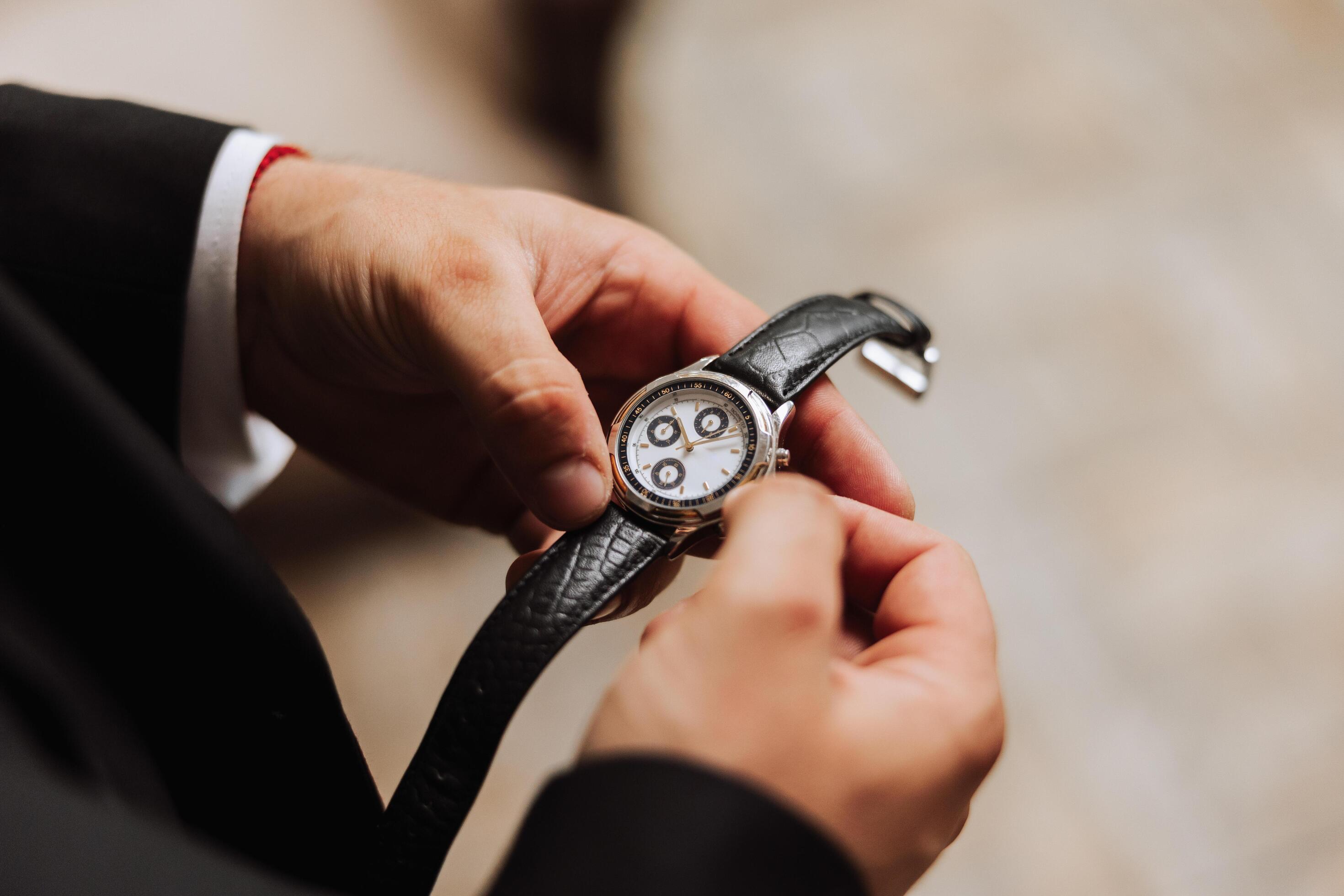 A man in a business suit holds a wristwatch in his hands, a mug of coffee is on the table in his room Stock Free