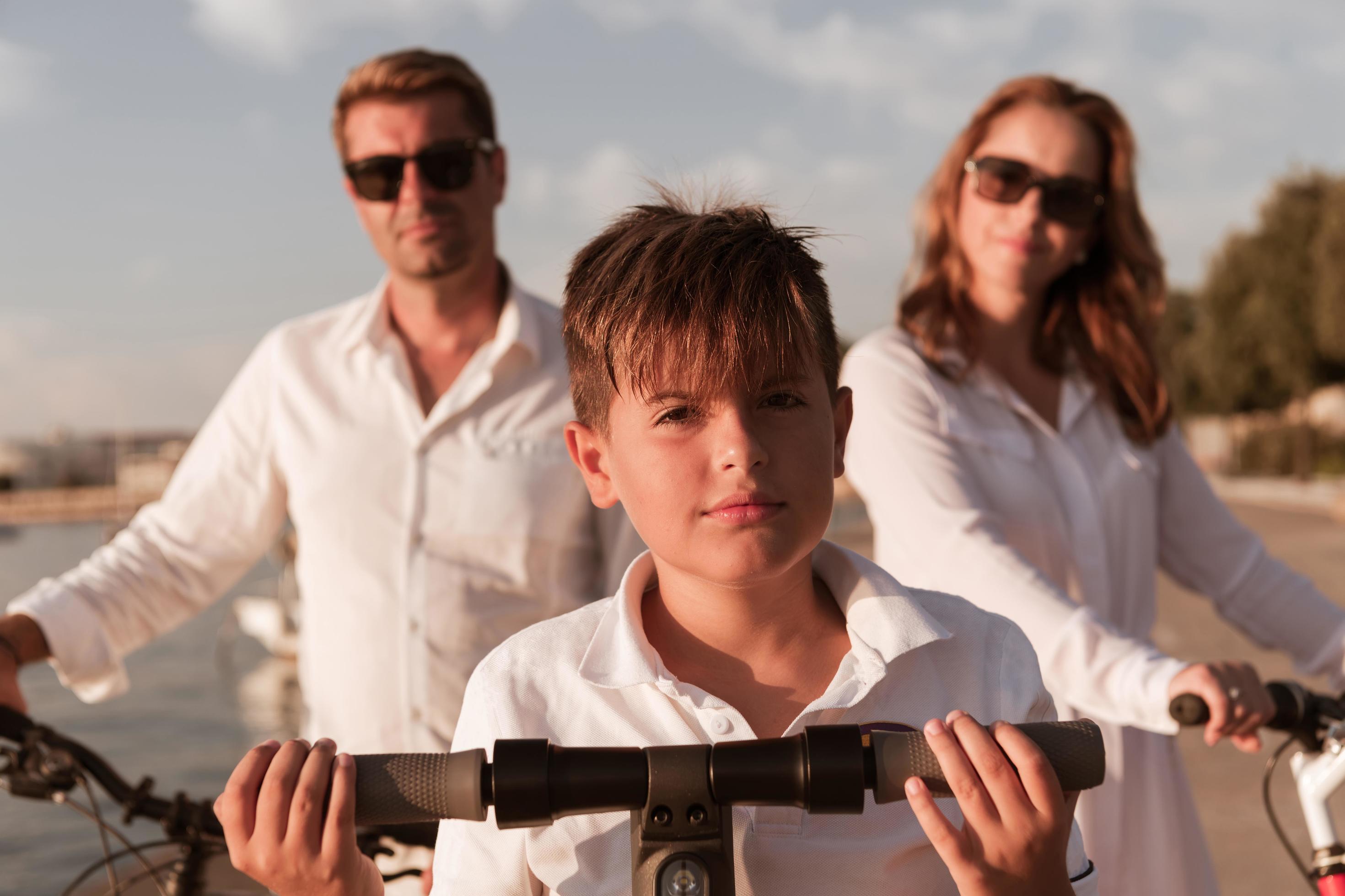 Happy family enjoying a beautiful morning by the sea together, parents riding a bike and their son riding an electric scooter. Selective focus Stock Free