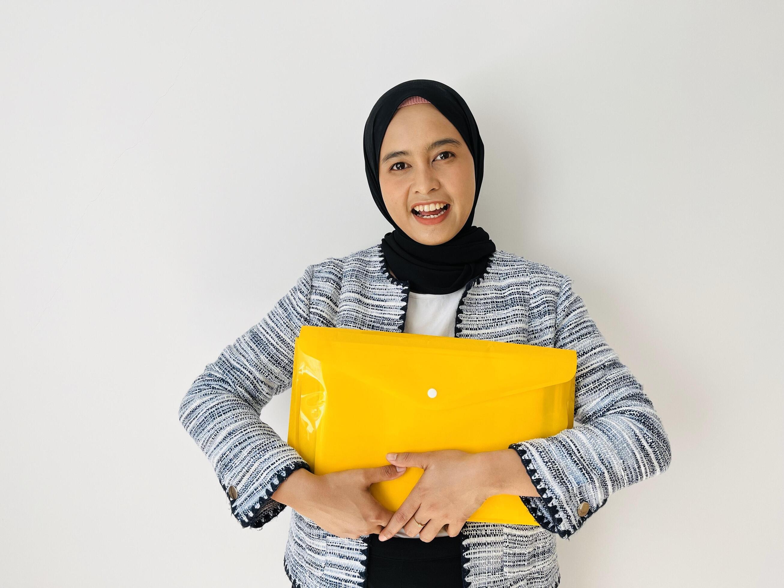 A young Asian woman holds a file folder in front of her chest happily. Business Women wear black headscarves and blazers Stock Free