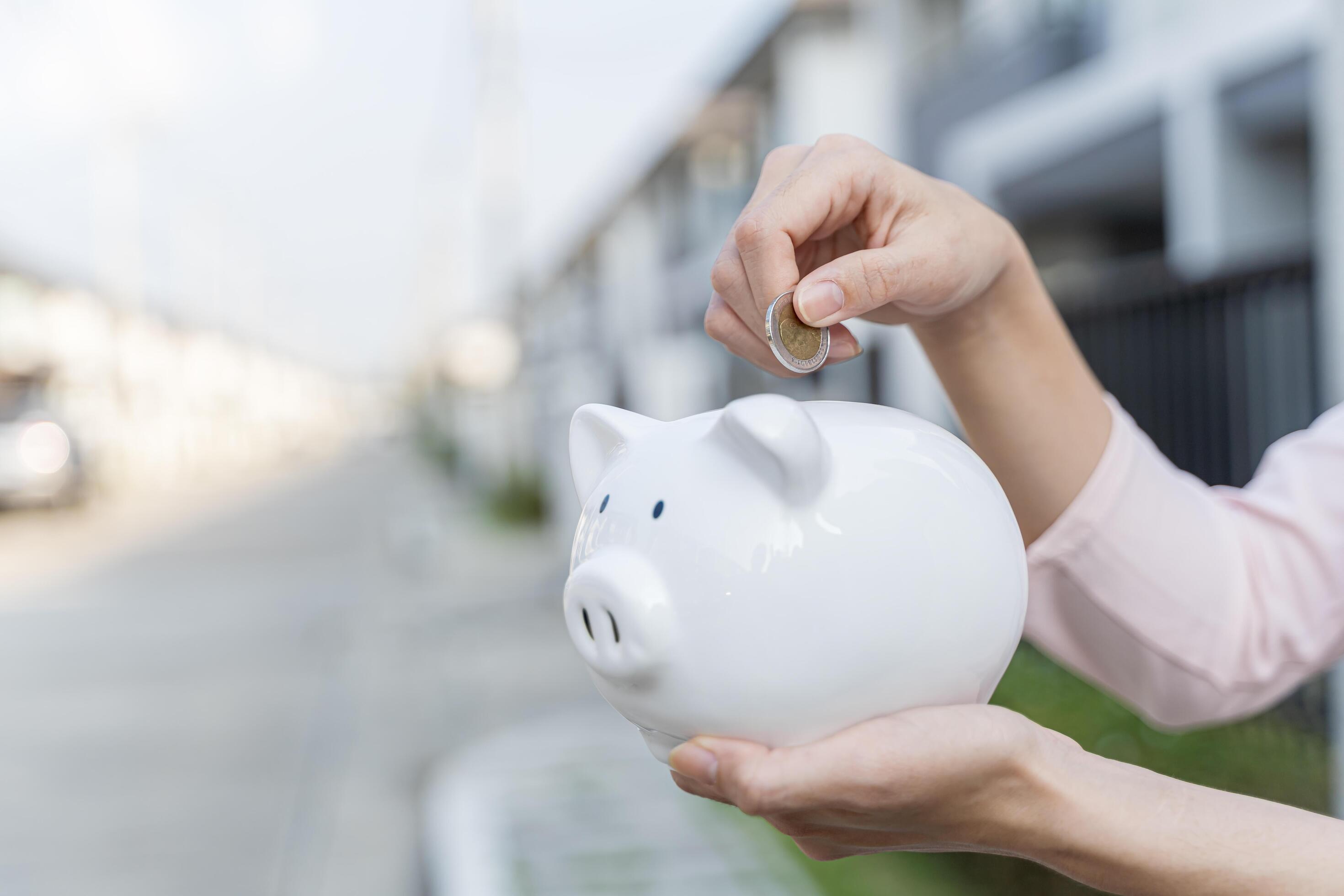 Woman hand hold a coin is save to the piggy bank with growing a interest and wooden home model for family in the public park Stock Free