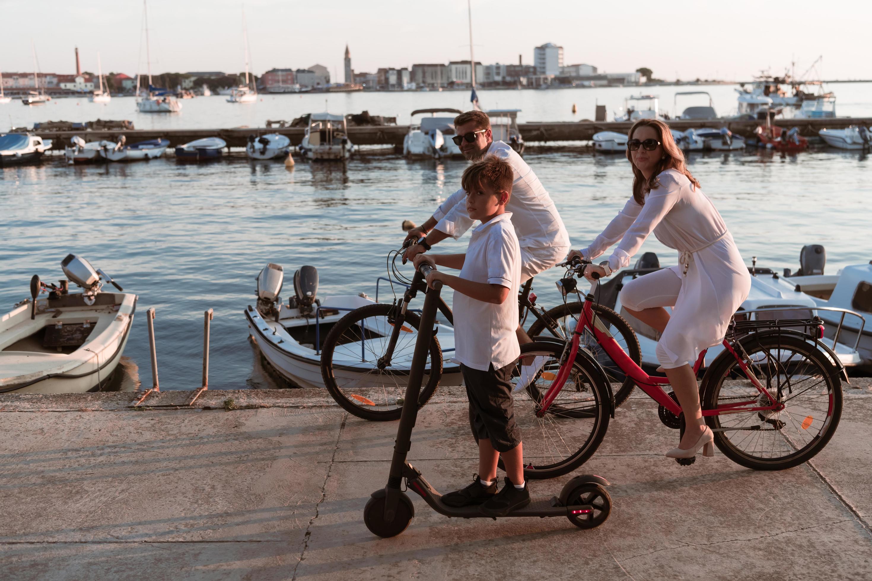 Happy family enjoying a beautiful morning by the sea together, parents riding a bike and their son riding an electric scooter. Selective focus Stock Free