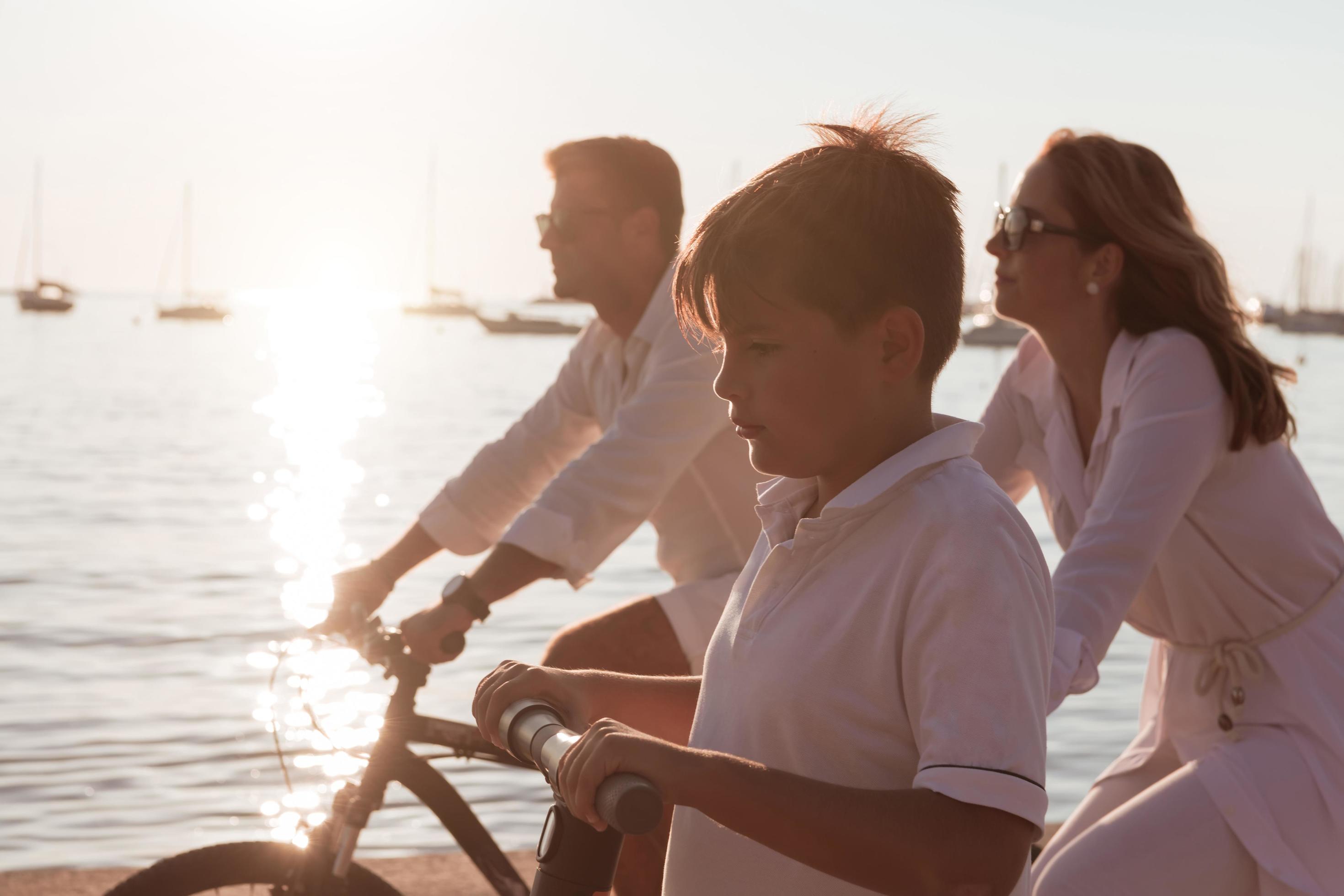 Happy family enjoying a beautiful morning by the sea together, parents riding a bike and their son riding an electric scooter. Selective focus Stock Free