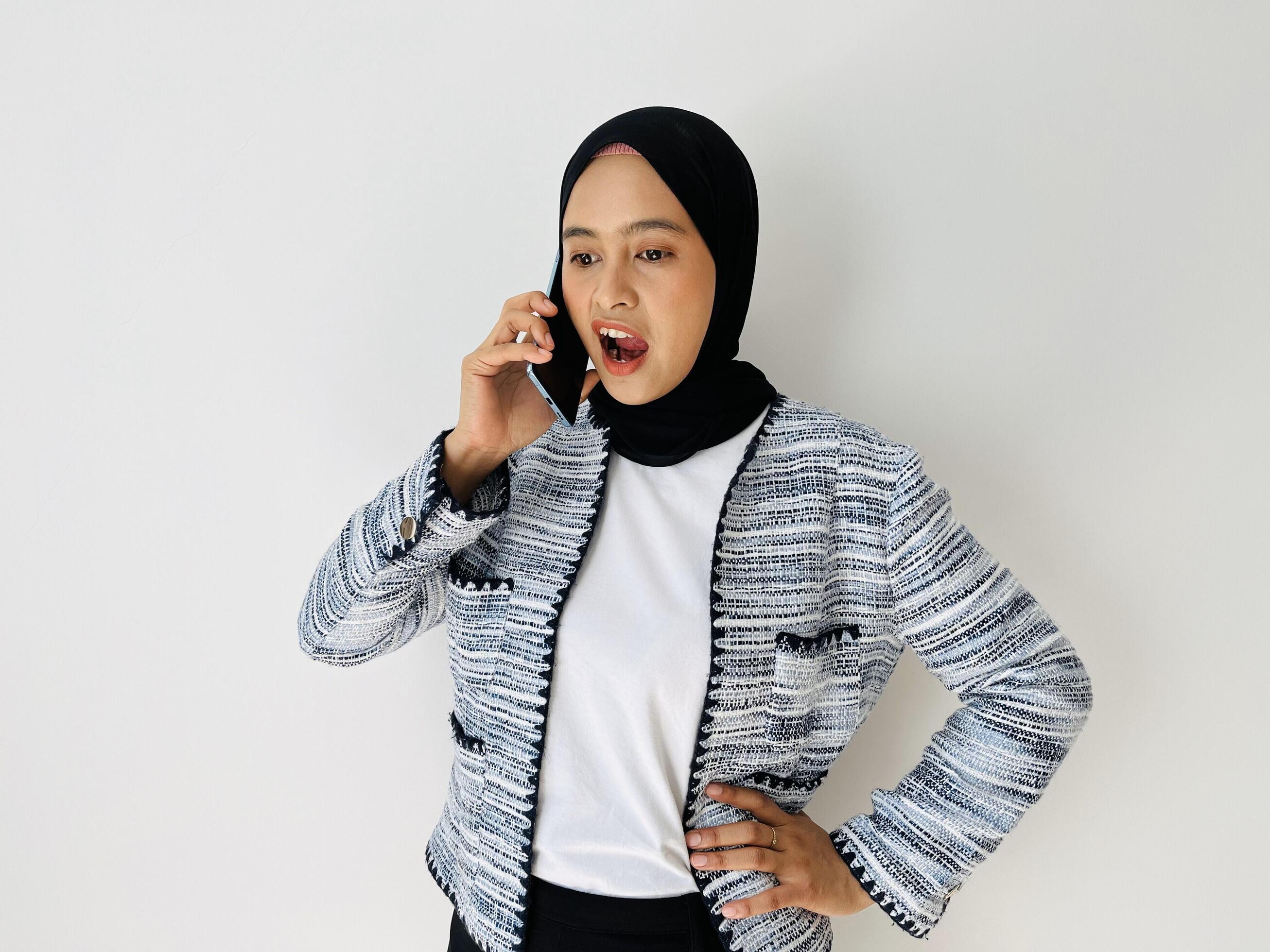 Angry young Asian woman using mobile phone having call. Standing female wearing black hijab and tweed blazer isolated on white background. Stock Free