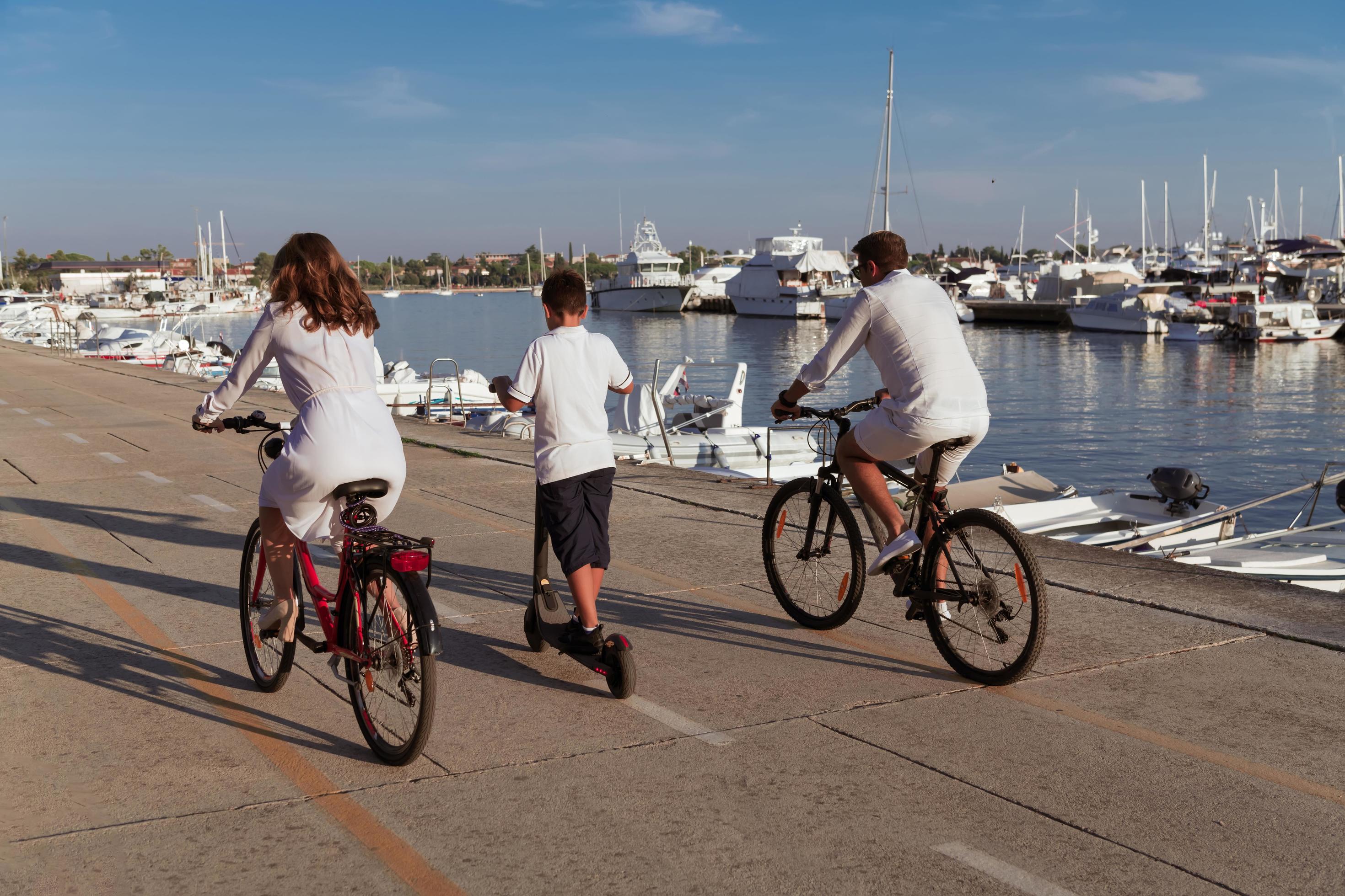 Happy family enjoying a beautiful morning by the sea together, parents riding a bike and their son riding an electric scooter. Selective focus Stock Free