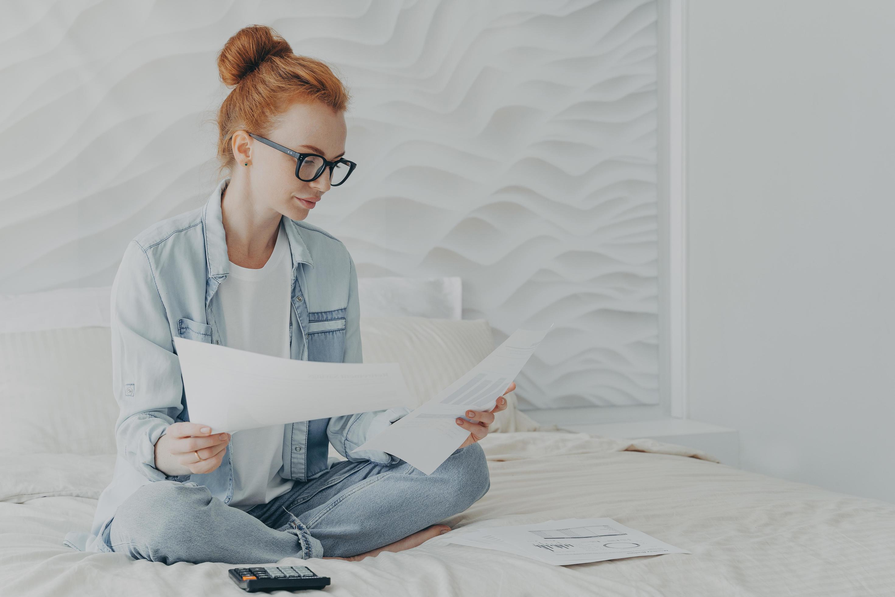 Young concentrated woman housewife in spectacles managing family budget while sitting on bed at home Stock Free