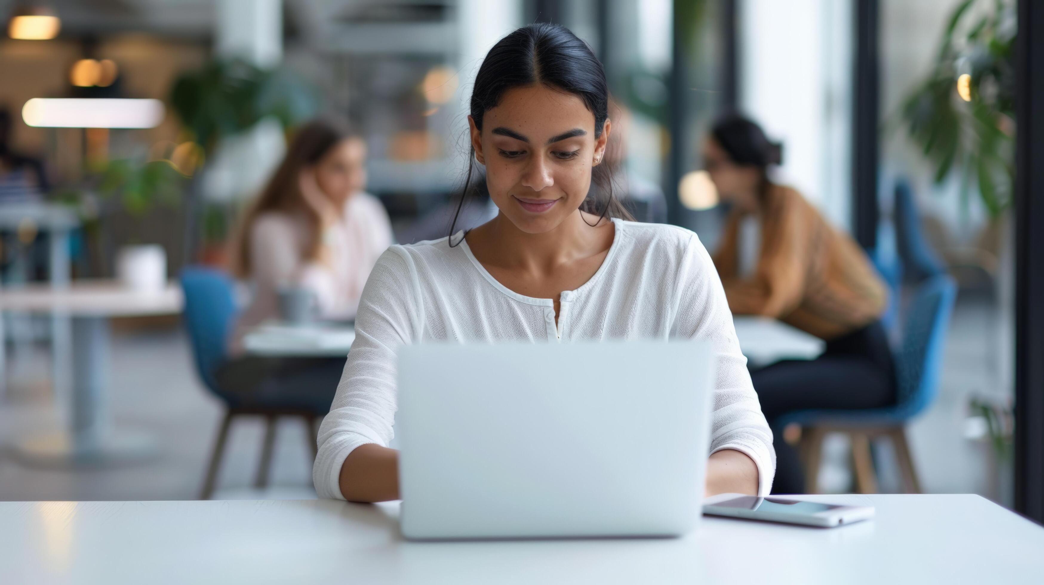 A focused young professional woman using a laptop in a modern office, working on her tasks with colleagues in the background Stock Free