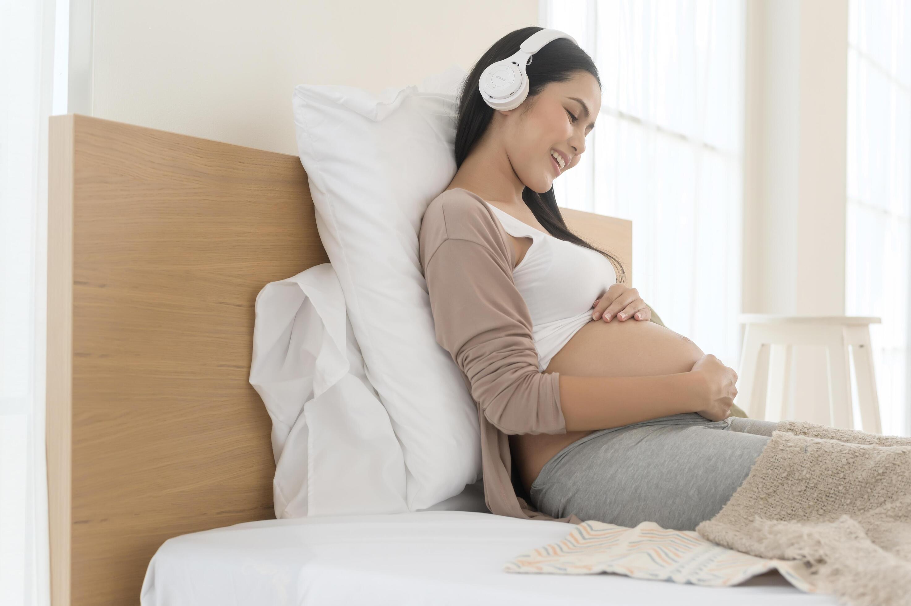 Happy pregnant woman with headphones listening to mozart music and lying on bed, pregnancy concept Stock Free