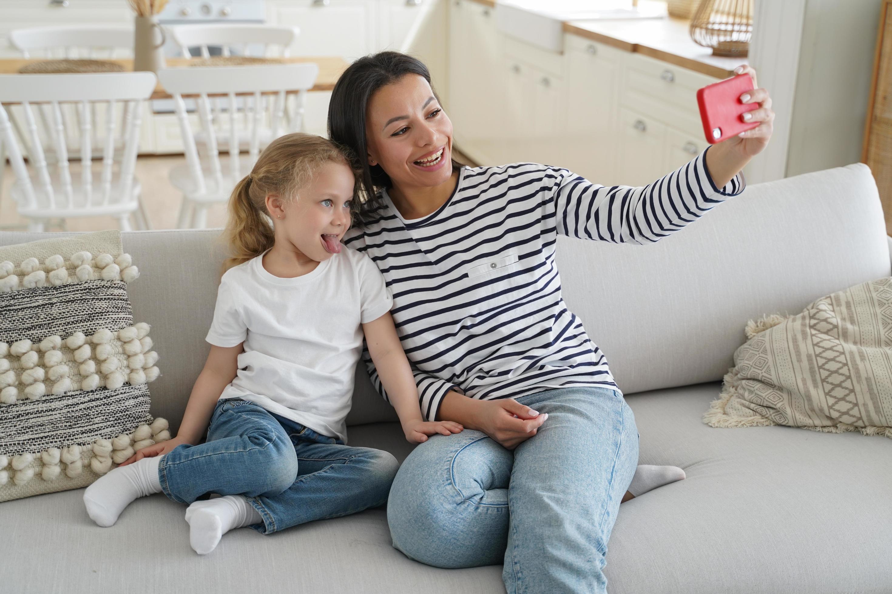 Mother, adopted little daughter, showing tongue, take family selfie together, using phone on couch Stock Free