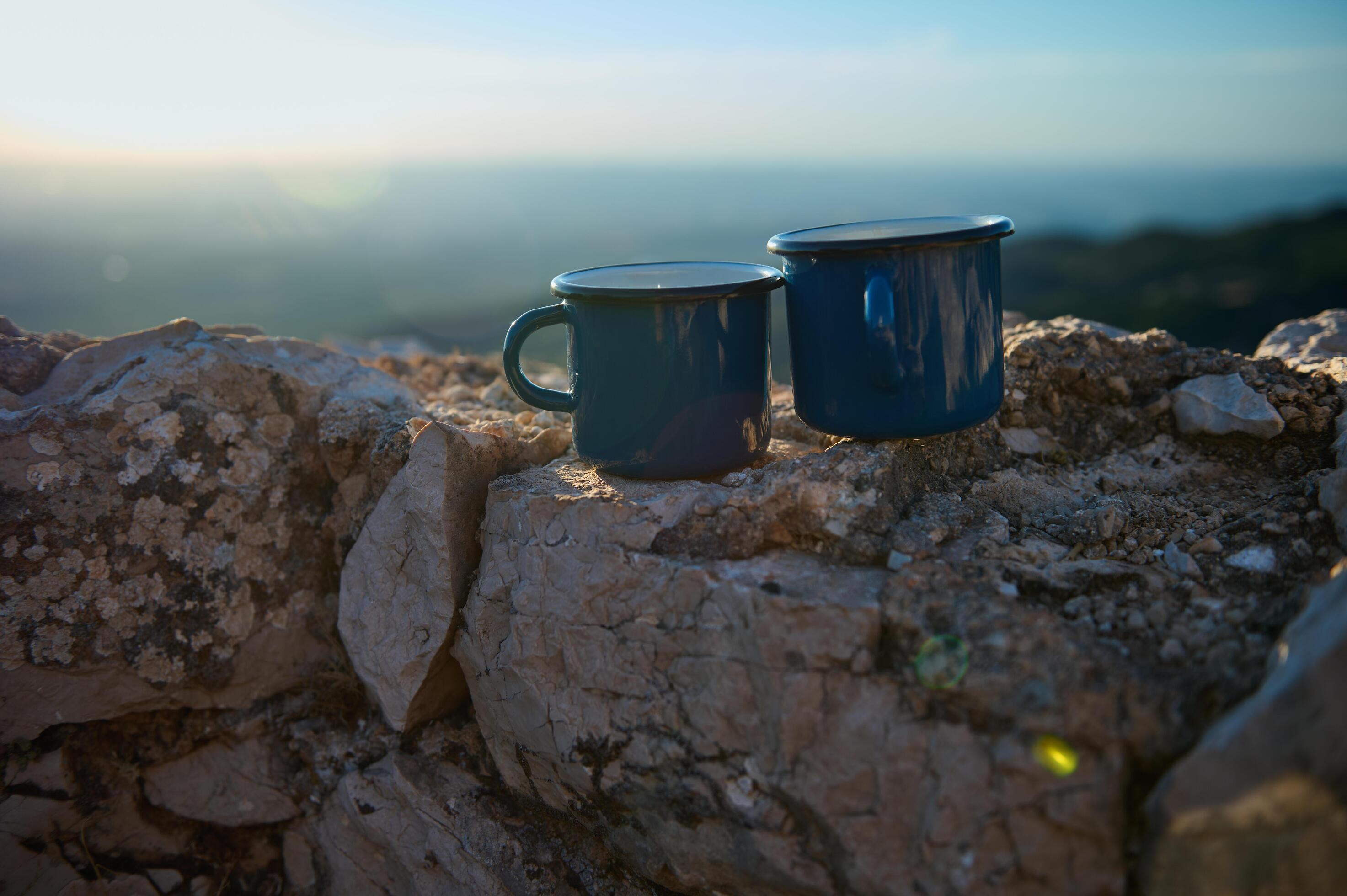 Two blue enamel mugs on rocky ground with a scenic background at sunrise Stock Free