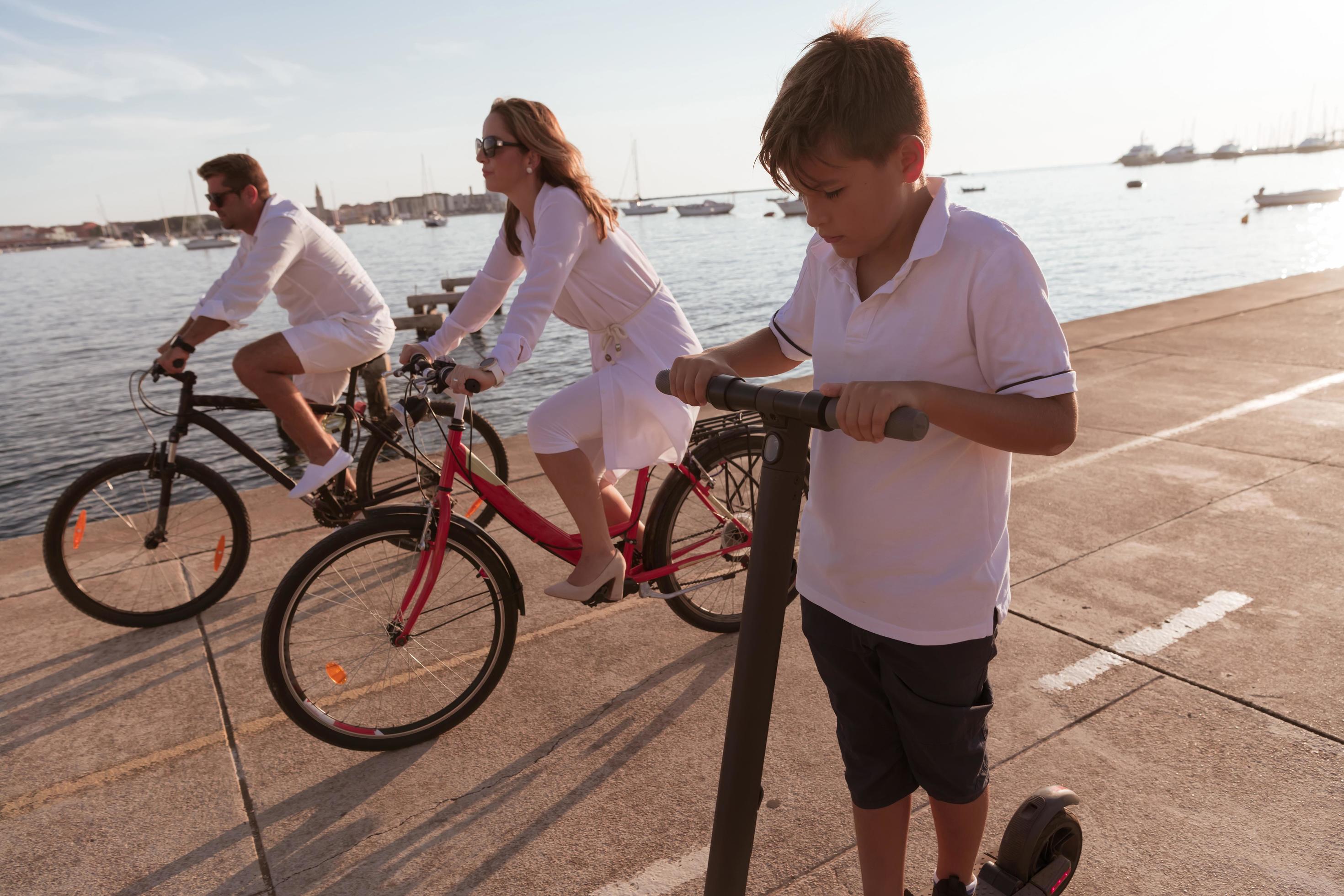 Happy family enjoying a beautiful morning by the sea together, parents riding a bike and their son riding an electric scooter. Selective focus Stock Free