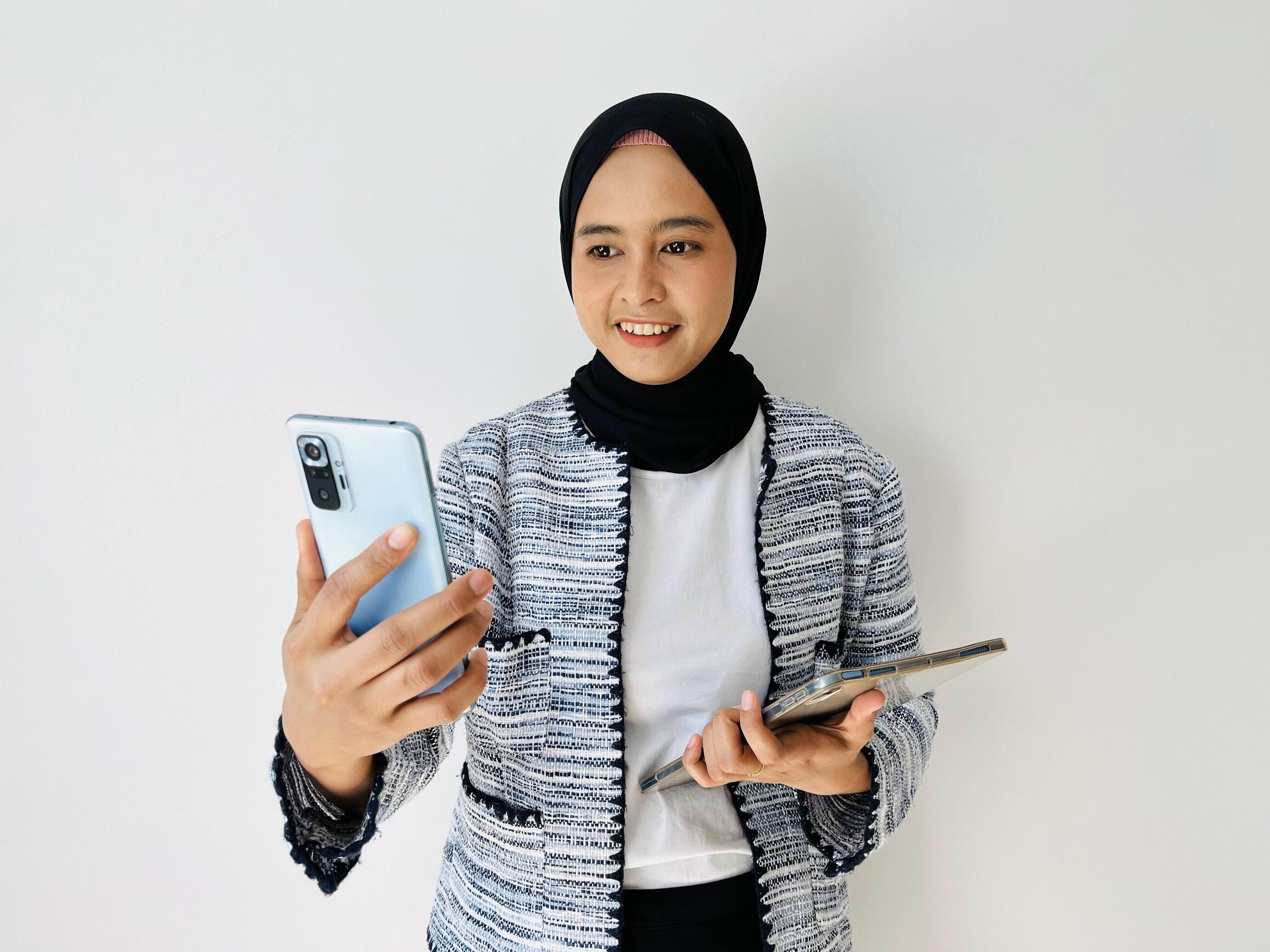 A young Asian woman is making a call for an online meeting using a mobile phone and holding a mobile tablet Stock Free