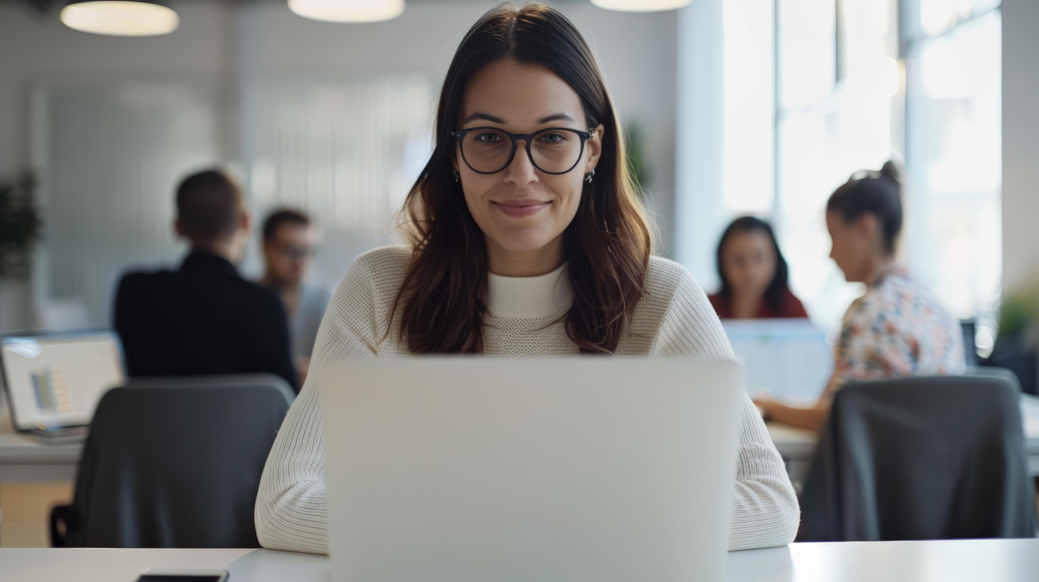 Young professional woman working on laptop in modern office setting with colleagues in the background, focused and productive Stock Free