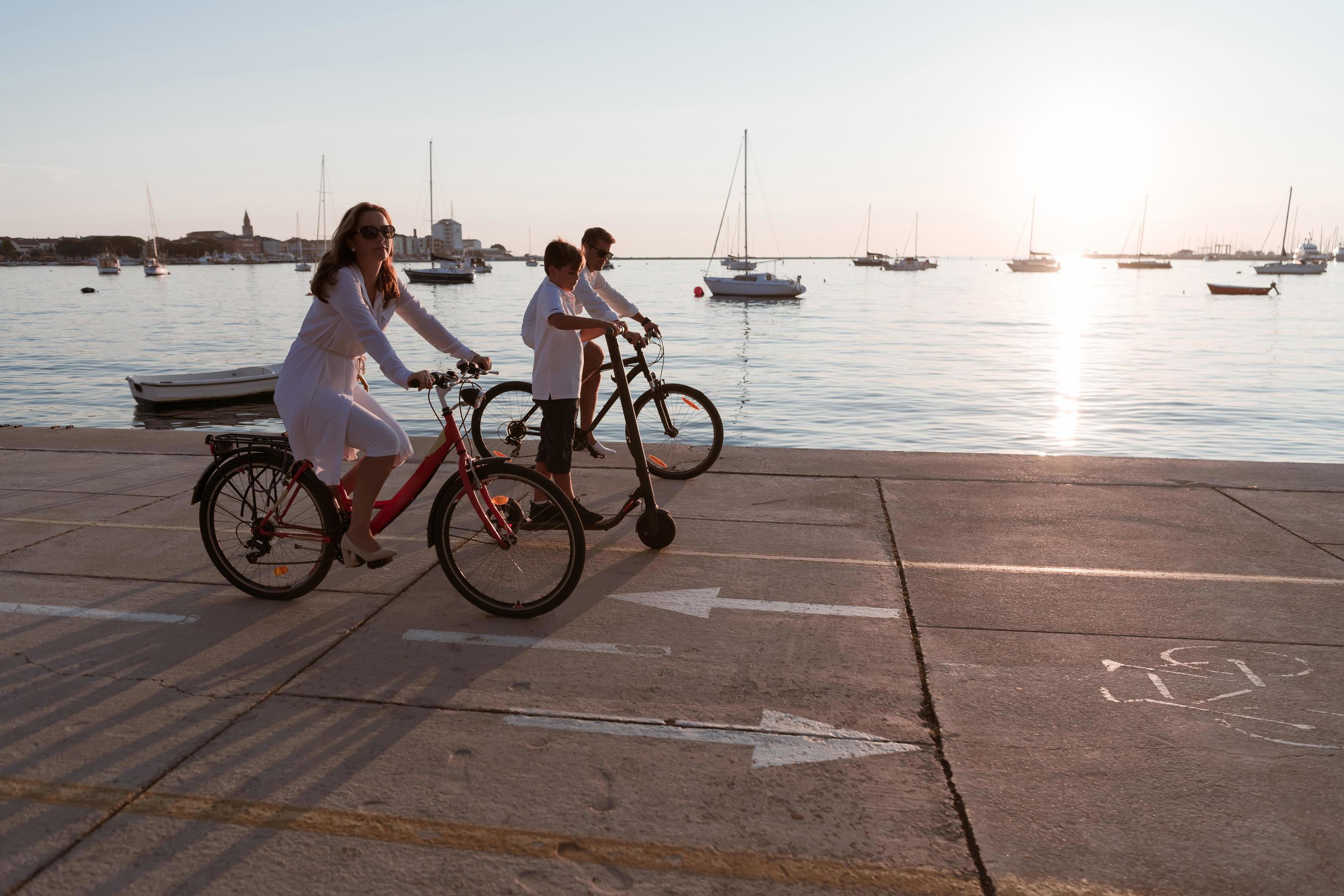 Happy family enjoying a beautiful morning by the sea together, parents riding a bike and their son riding an electric scooter. Selective focus Stock Free