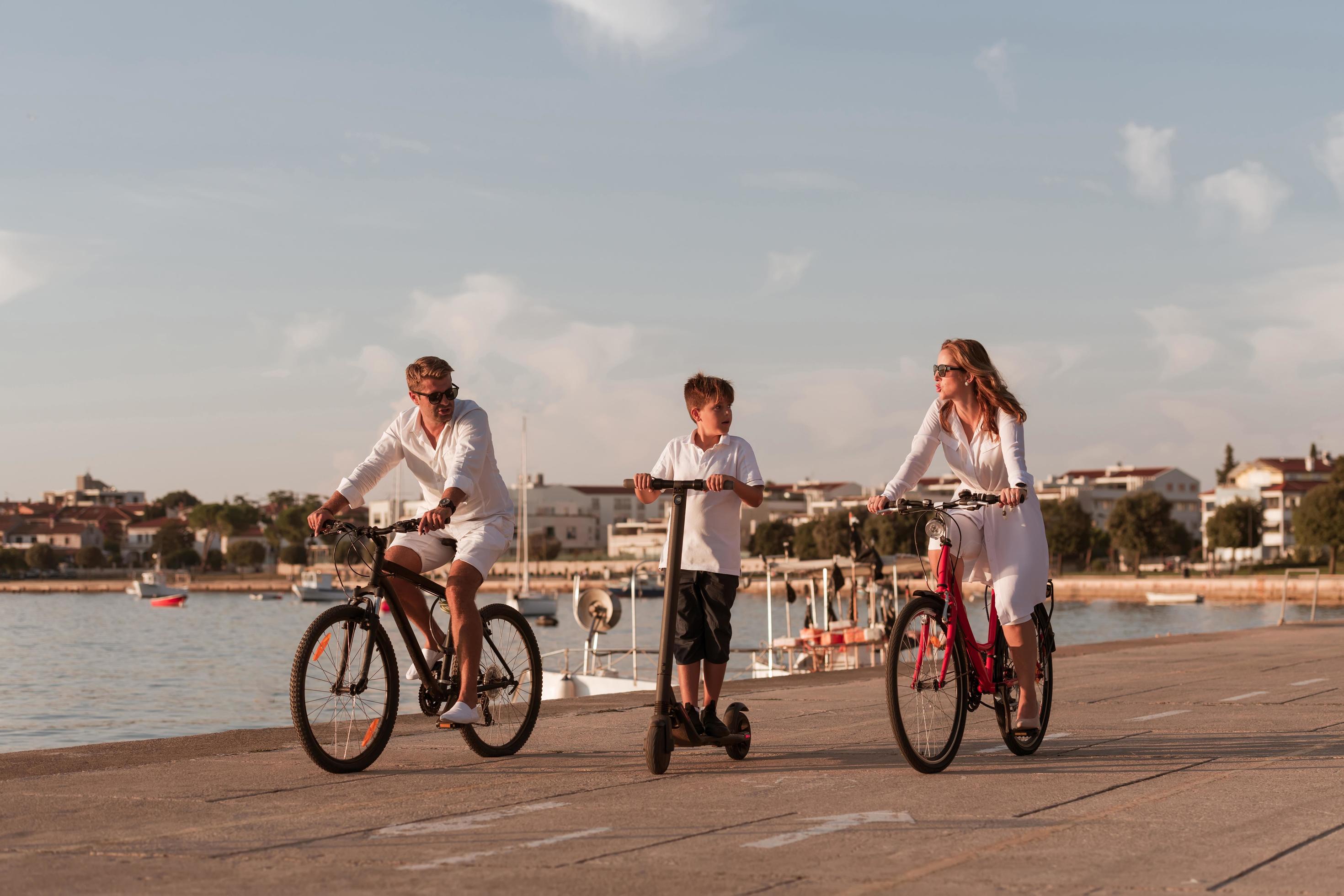 Happy family enjoying a beautiful morning by the sea together, parents riding a bike and their son riding an electric scooter. Selective focus Stock Free