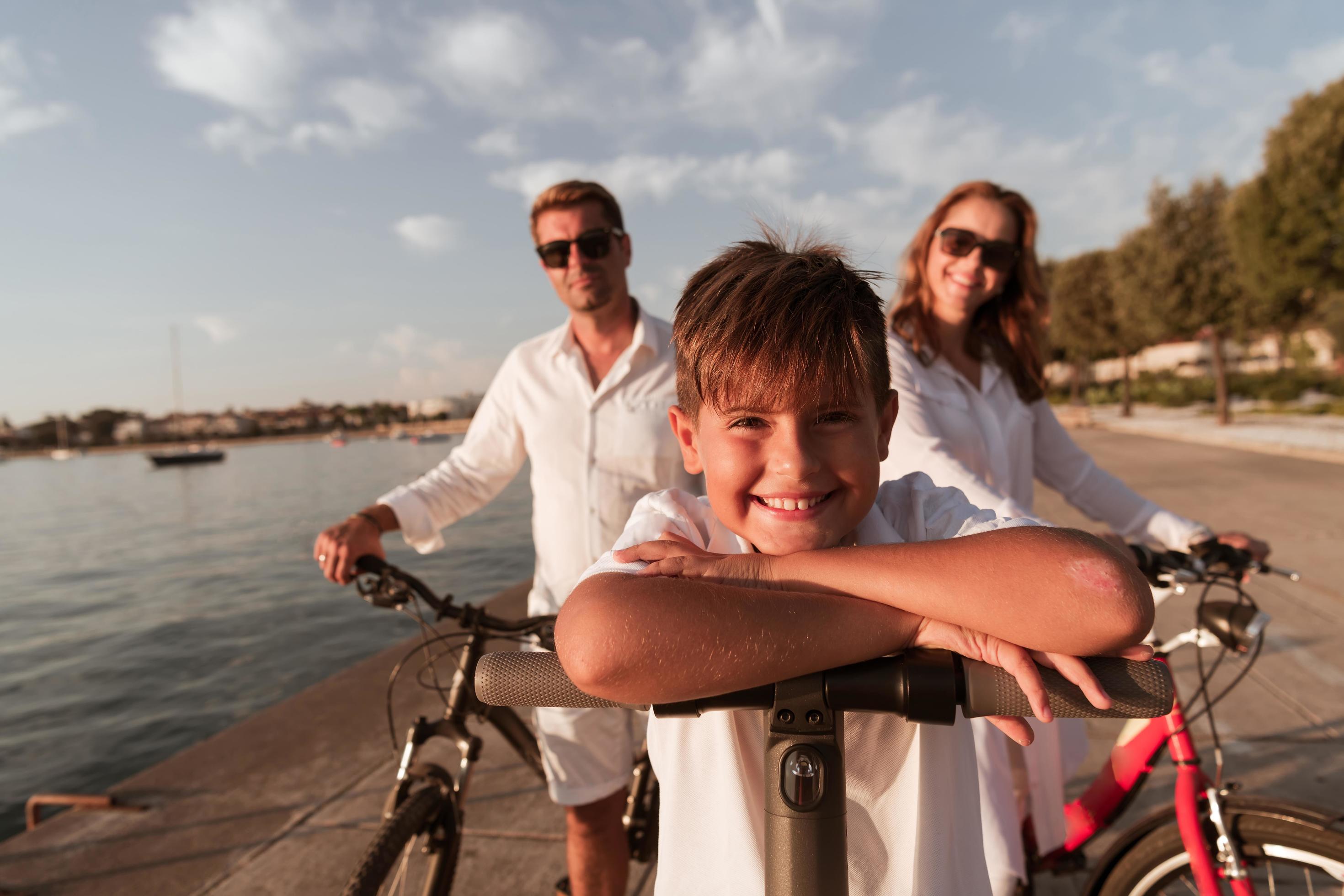 Happy family enjoying a beautiful morning by the sea together, parents riding a bike and their son riding an electric scooter. Selective focus Stock Free