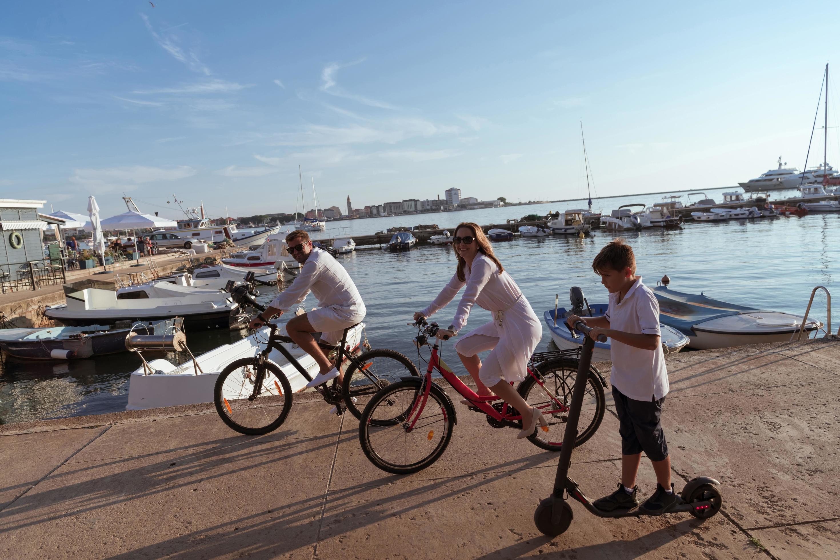 Happy family enjoying a beautiful morning by the sea together, parents riding a bike and their son riding an electric scooter. Selective focus Stock Free