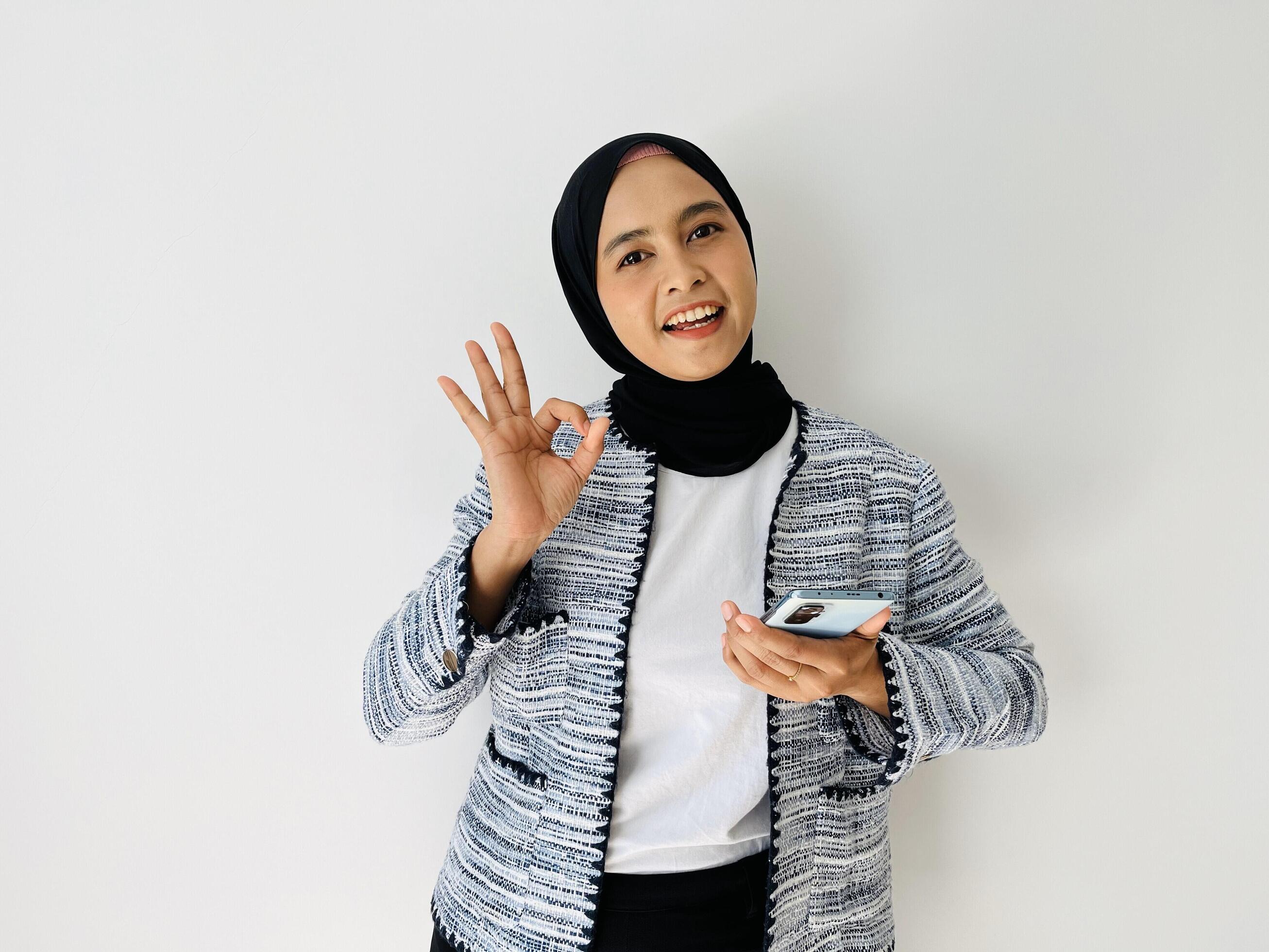 Portrait of smiling Asian Muslim woman OK gesture with the smartphone wearing black hijab and tweed blazer over white background Stock Free