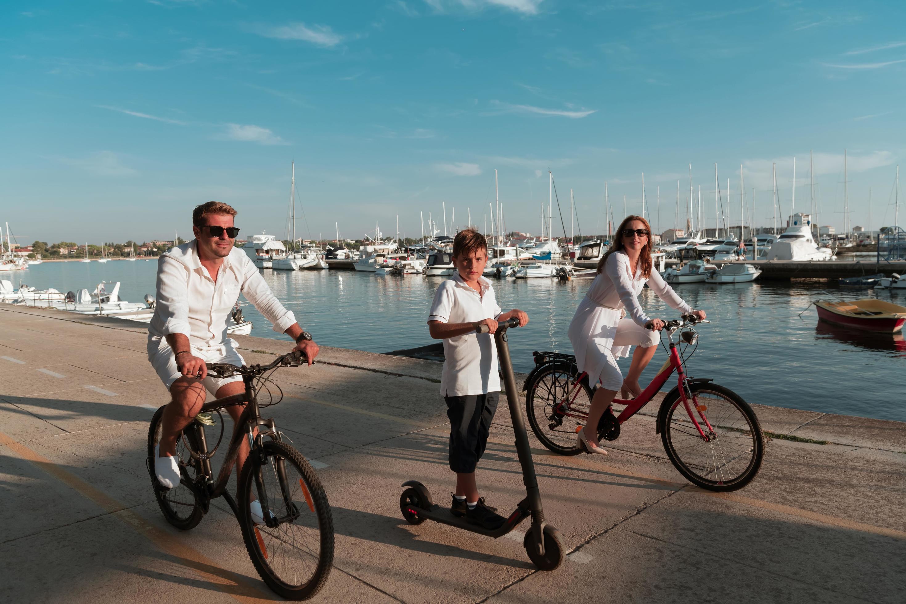 Happy family enjoying a beautiful morning by the sea together, parents riding a bike and their son riding an electric scooter. Selective focus Stock Free