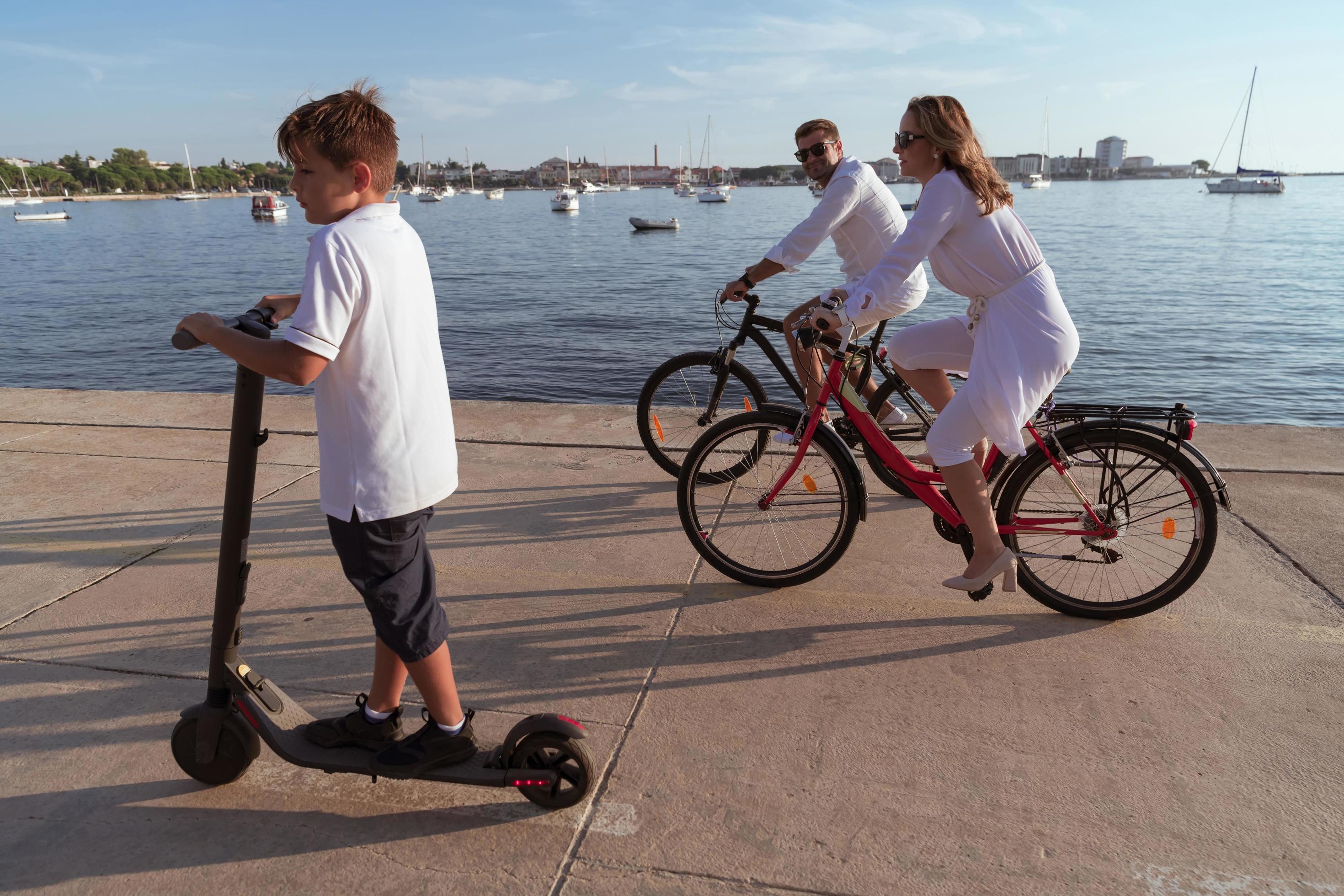 Happy family enjoying a beautiful morning by the sea together, parents riding a bike and their son riding an electric scooter. Selective focus Stock Free