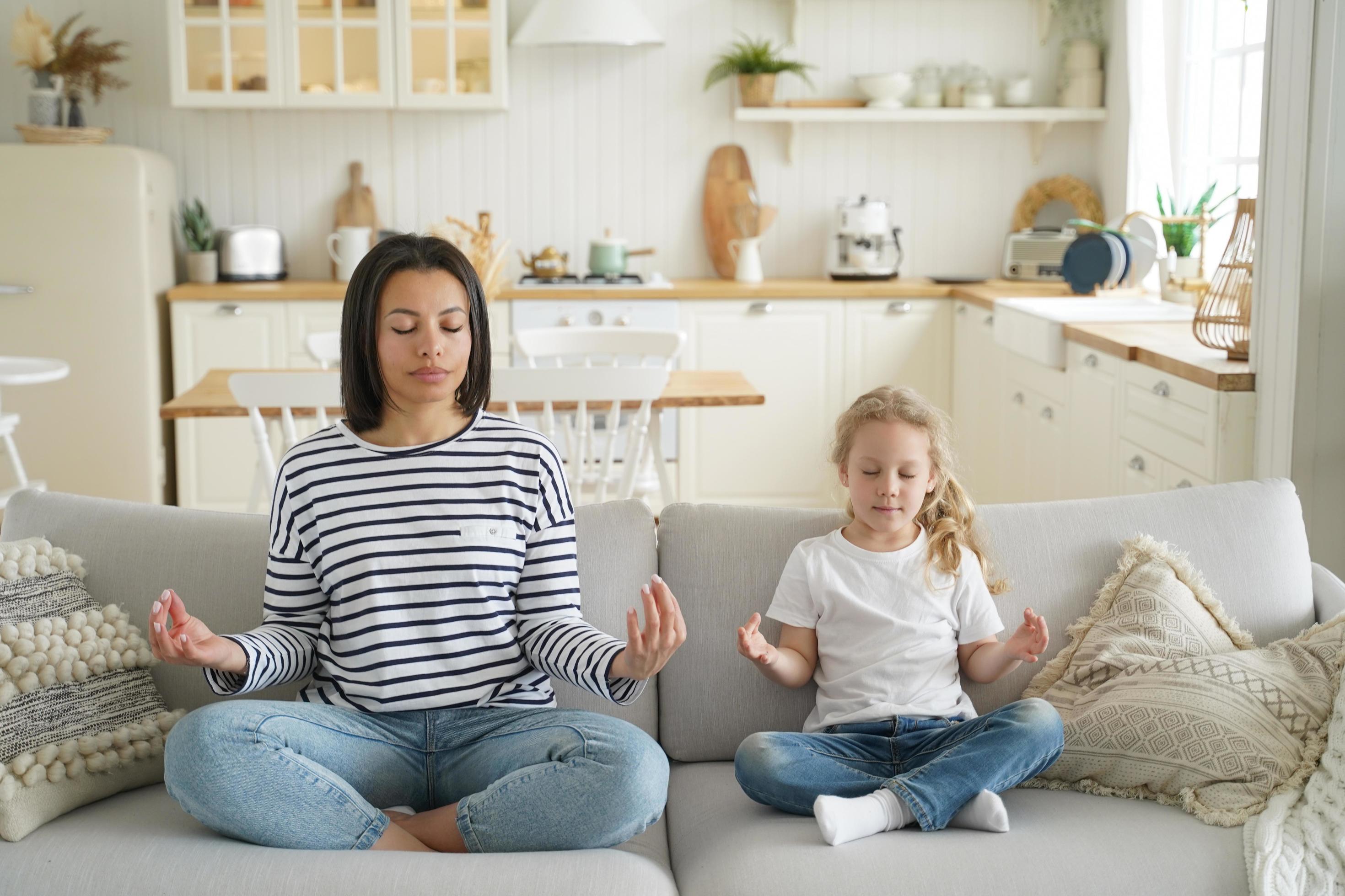 Mother, foster daughter practice yoga sitting on sofa at home. Healthy lifestyle, adoption of child Stock Free