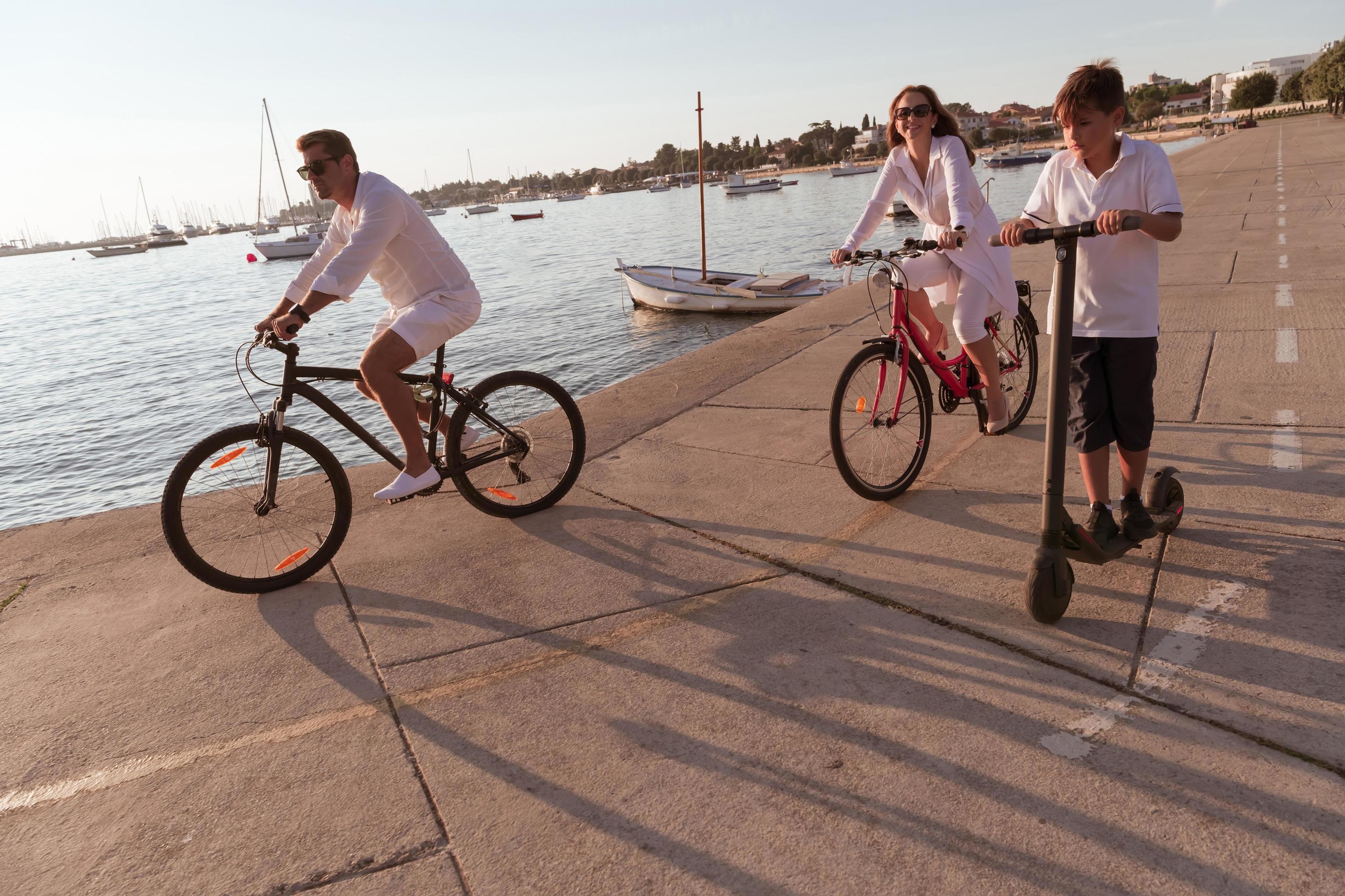 Happy family enjoying a beautiful morning by the sea together, parents riding a bike and their son riding an electric scooter. Selective focus Stock Free