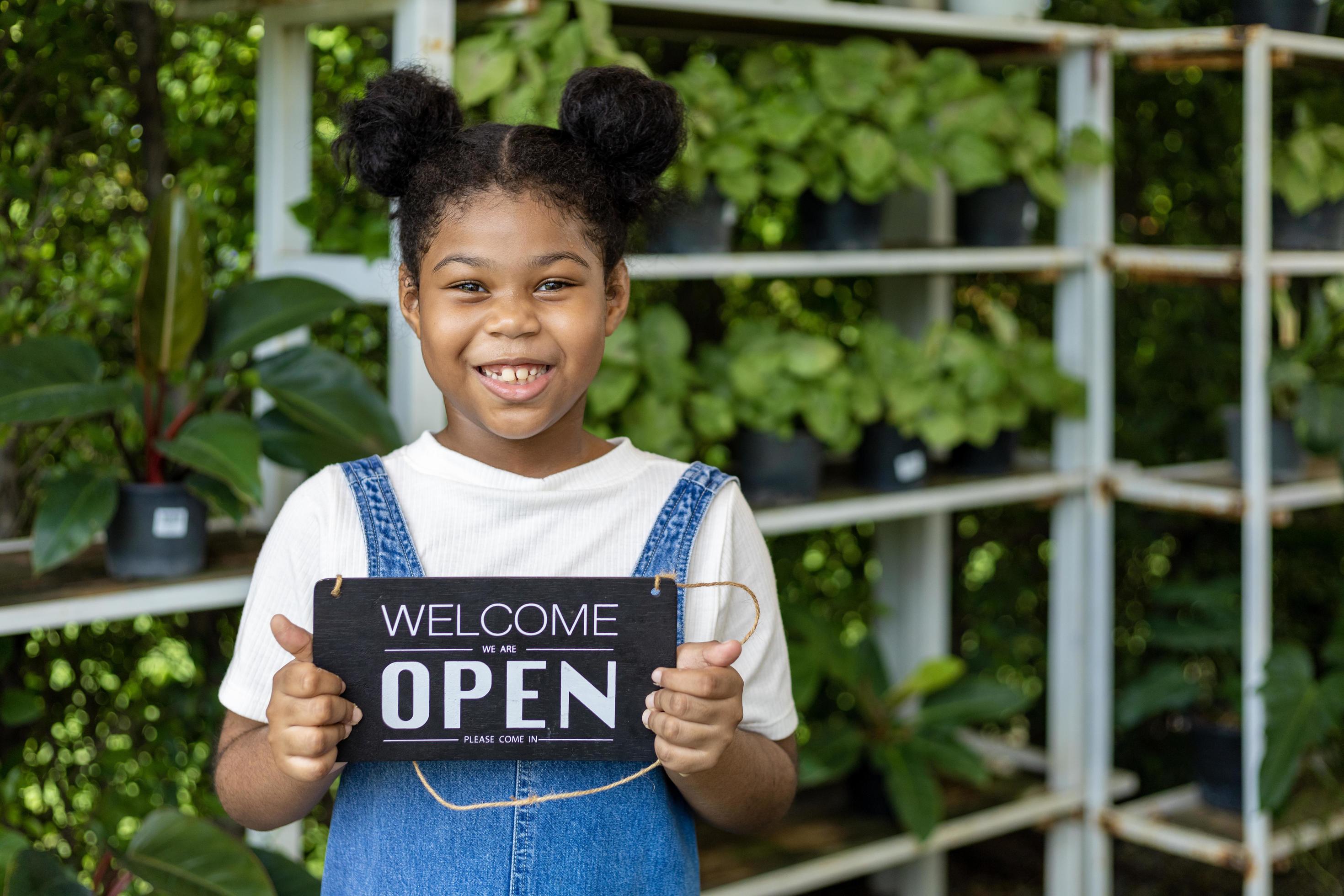 Portrait of African American girl holding welcome open sign for her family garden center business for nursery and houseplant shop Stock Free