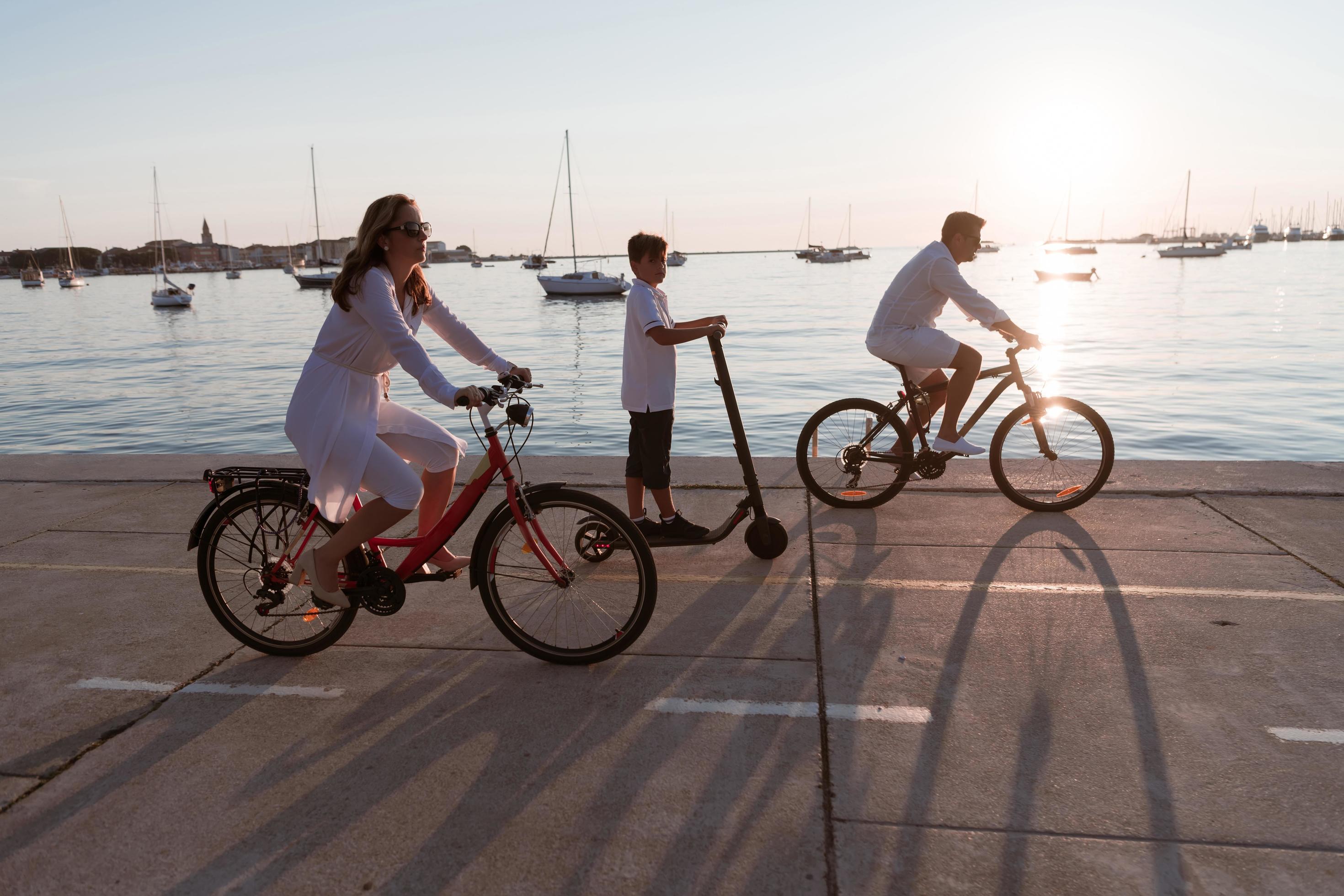 Happy family enjoying a beautiful morning by the sea together, parents riding a bike and their son riding an electric scooter. Selective focus Stock Free