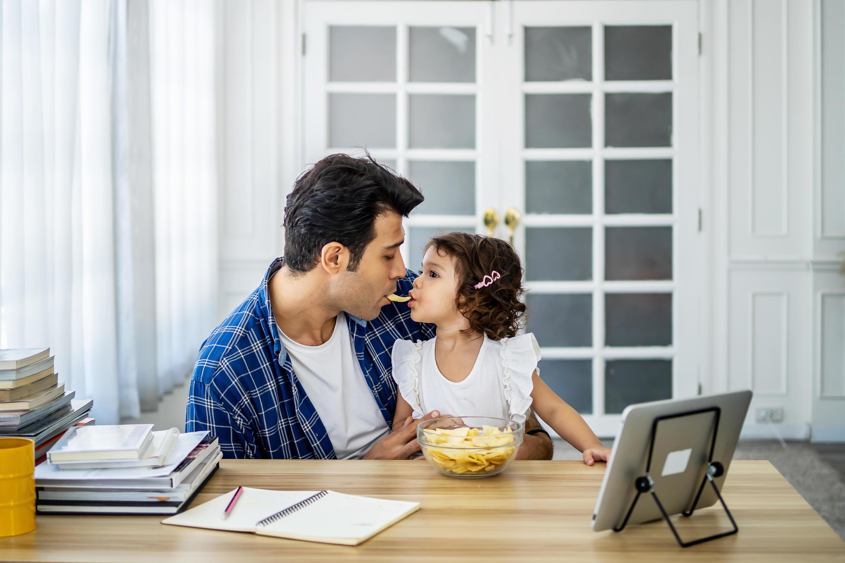 Caucasian family young father eating potato chips in mounth with little daughter together and watch cartoons educational online program Stock Free