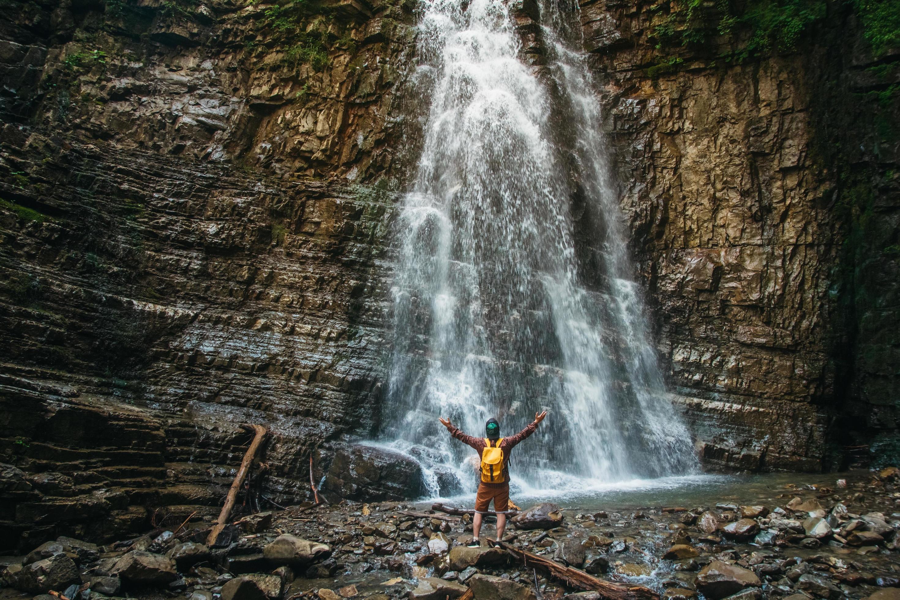 Traveler man with a yellow backpack standing on background of a waterfall. Travel lifestyle concept. Stock Free