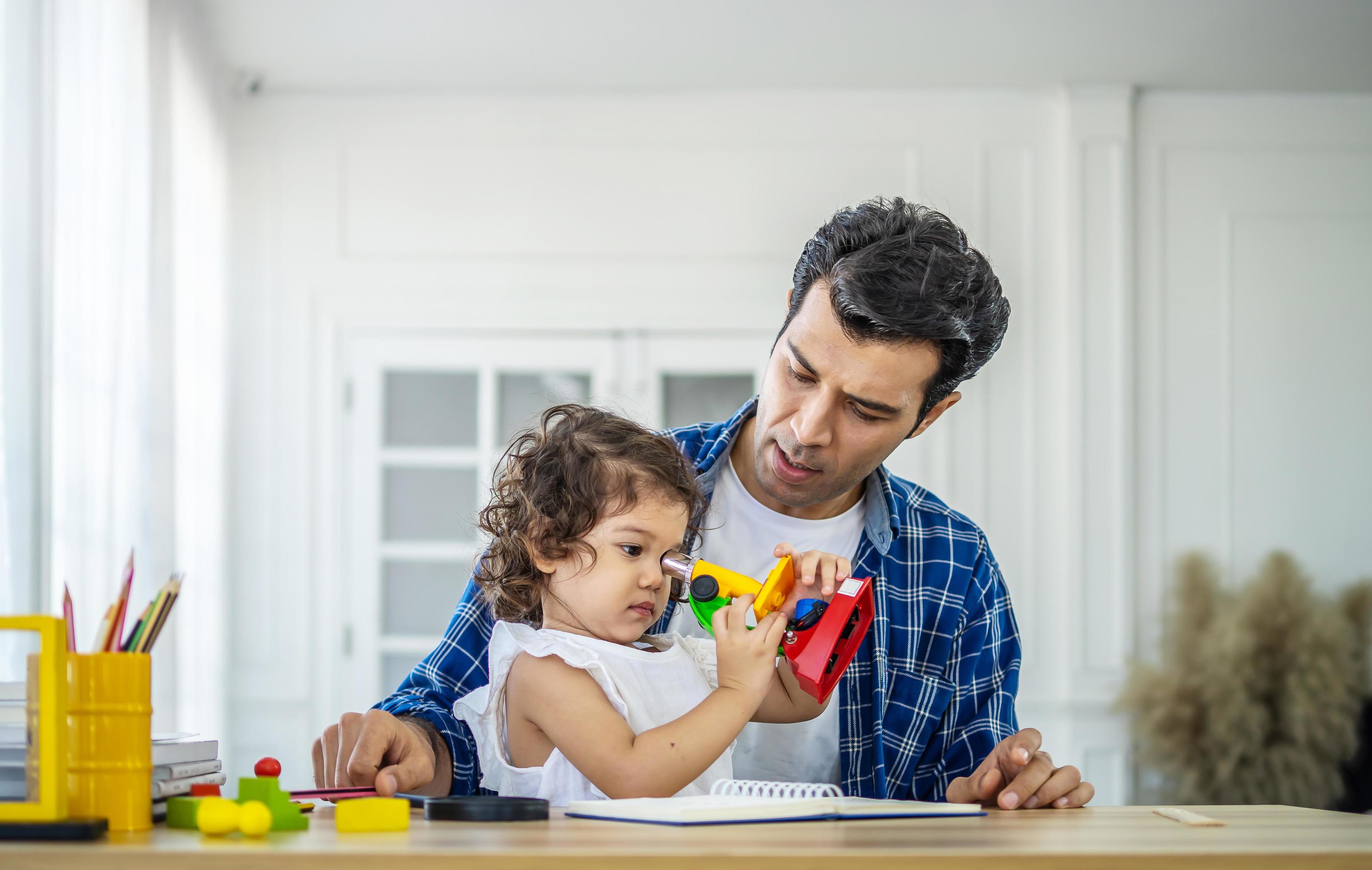 family, fatherhood and childhood concept – happy father and little daughter playing with toy microscope at table at home Stock Free