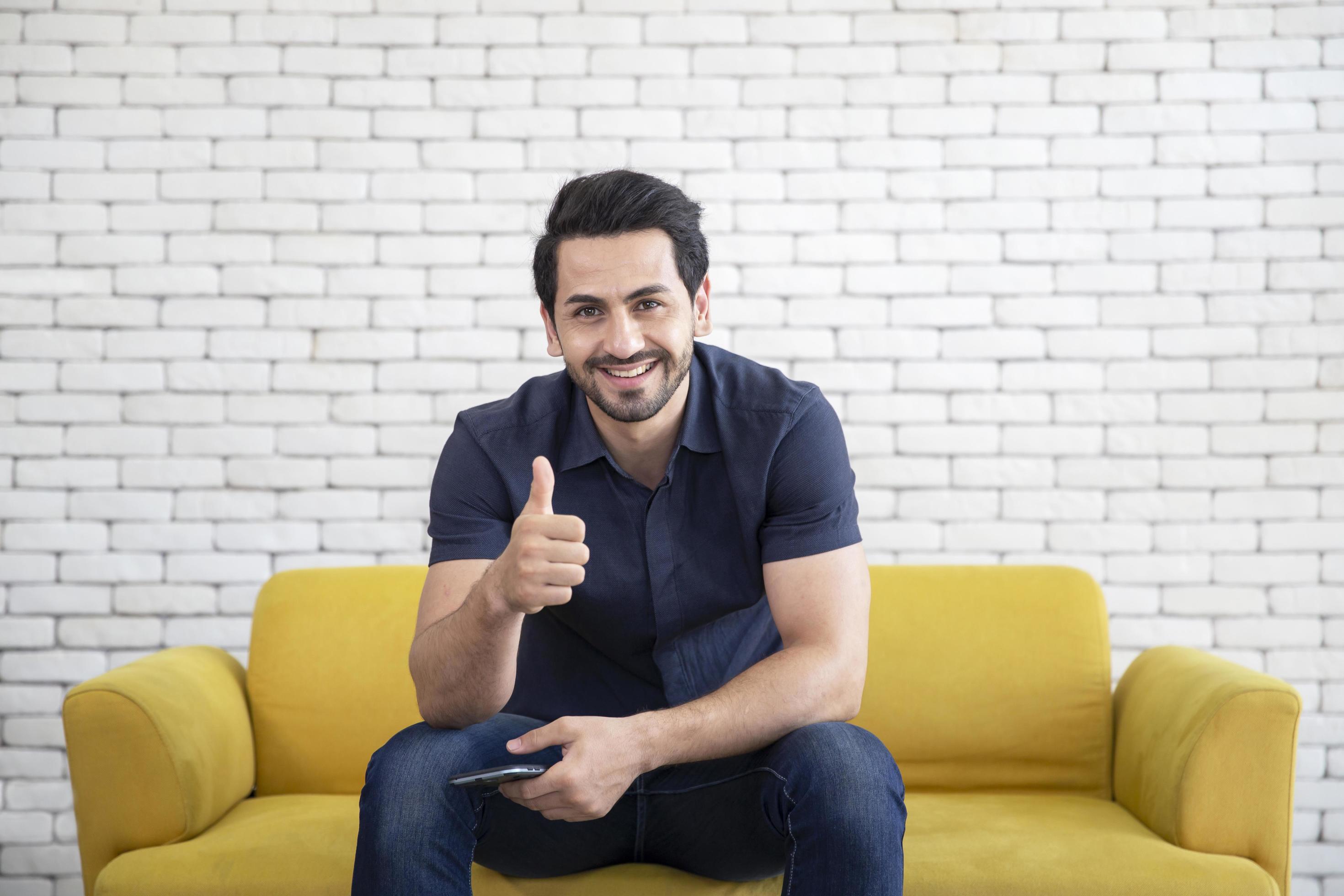 Lifestyle portrait of attractive handsome man with pleasant smile relaxing and sitting on yellow sofa at living room Stock Free