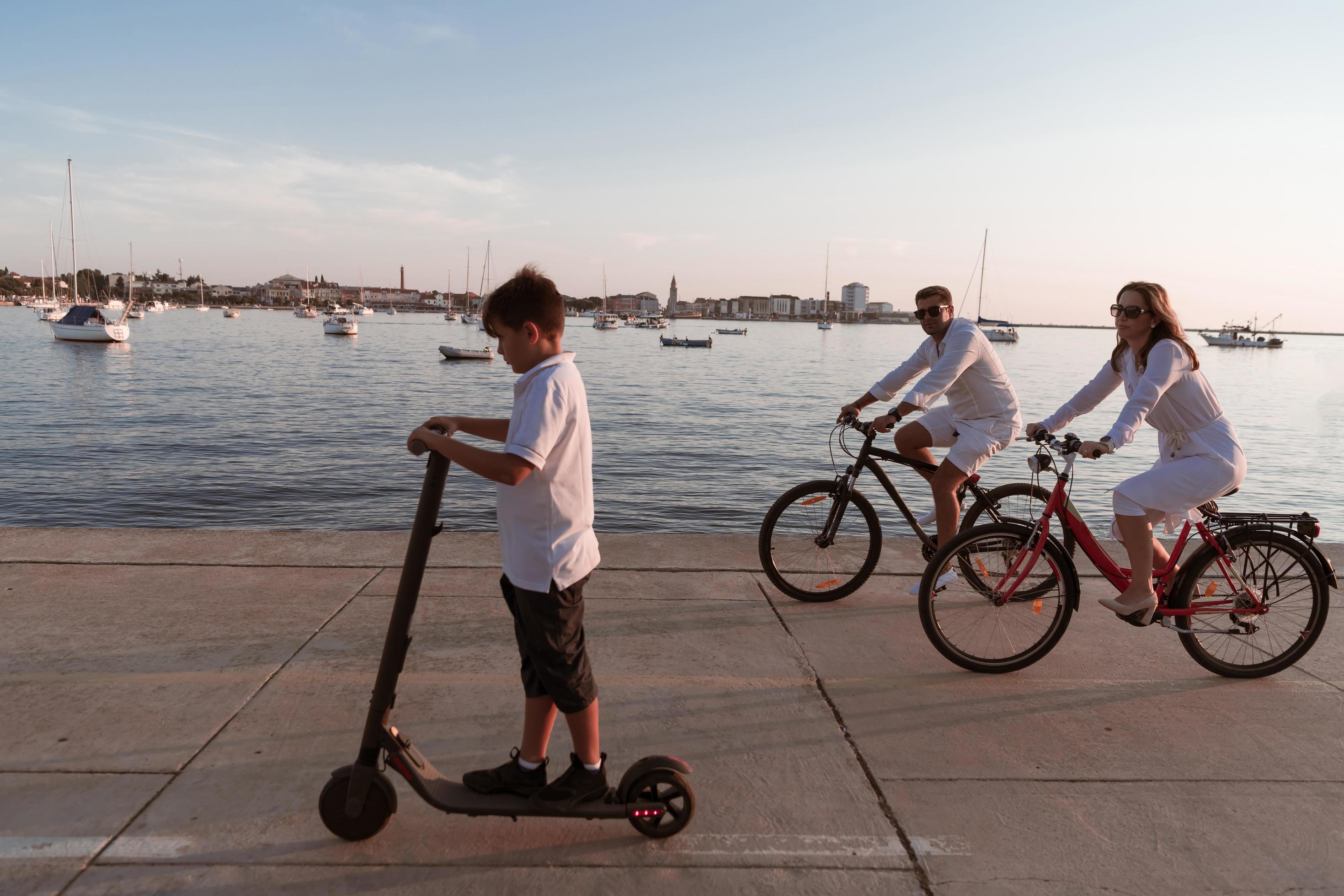 Happy family enjoying a beautiful morning by the sea together, parents riding a bike and their son riding an electric scooter. Selective focus Stock Free