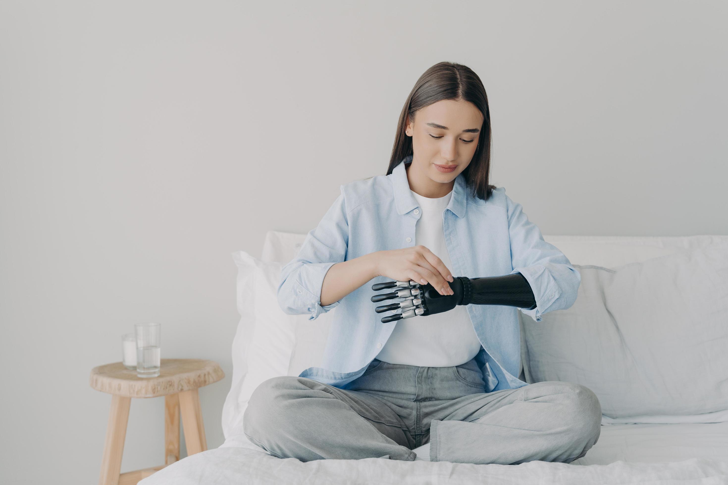 Disabled girl setting bionic arm prosthesis, sitting on bed. Lifestyle of people with disabilities Stock Free