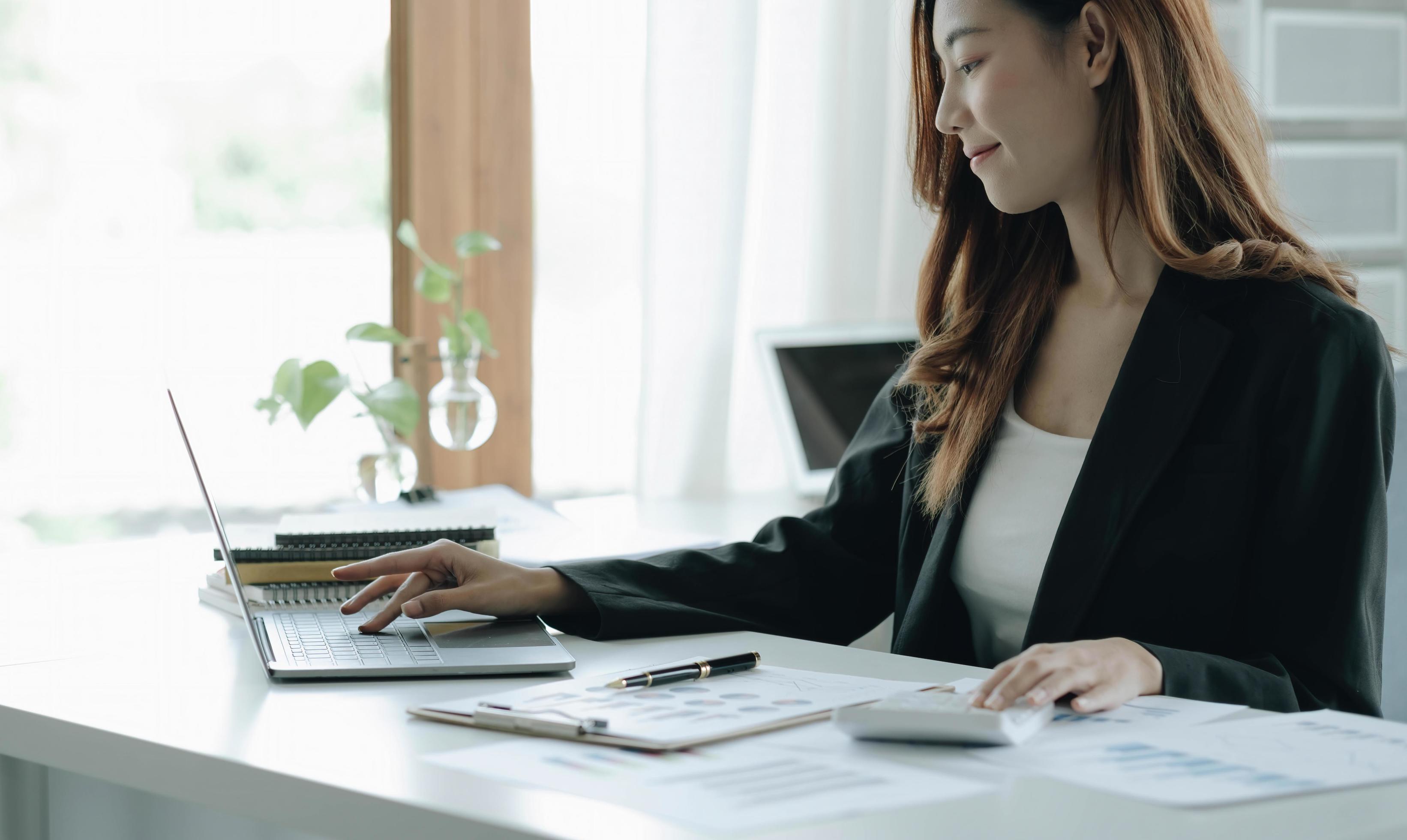 Beautiful Asian woman sitting in office using laptop and calculator. Happy business woman sitting at a desk in an office with a tablet computer. Stock Free