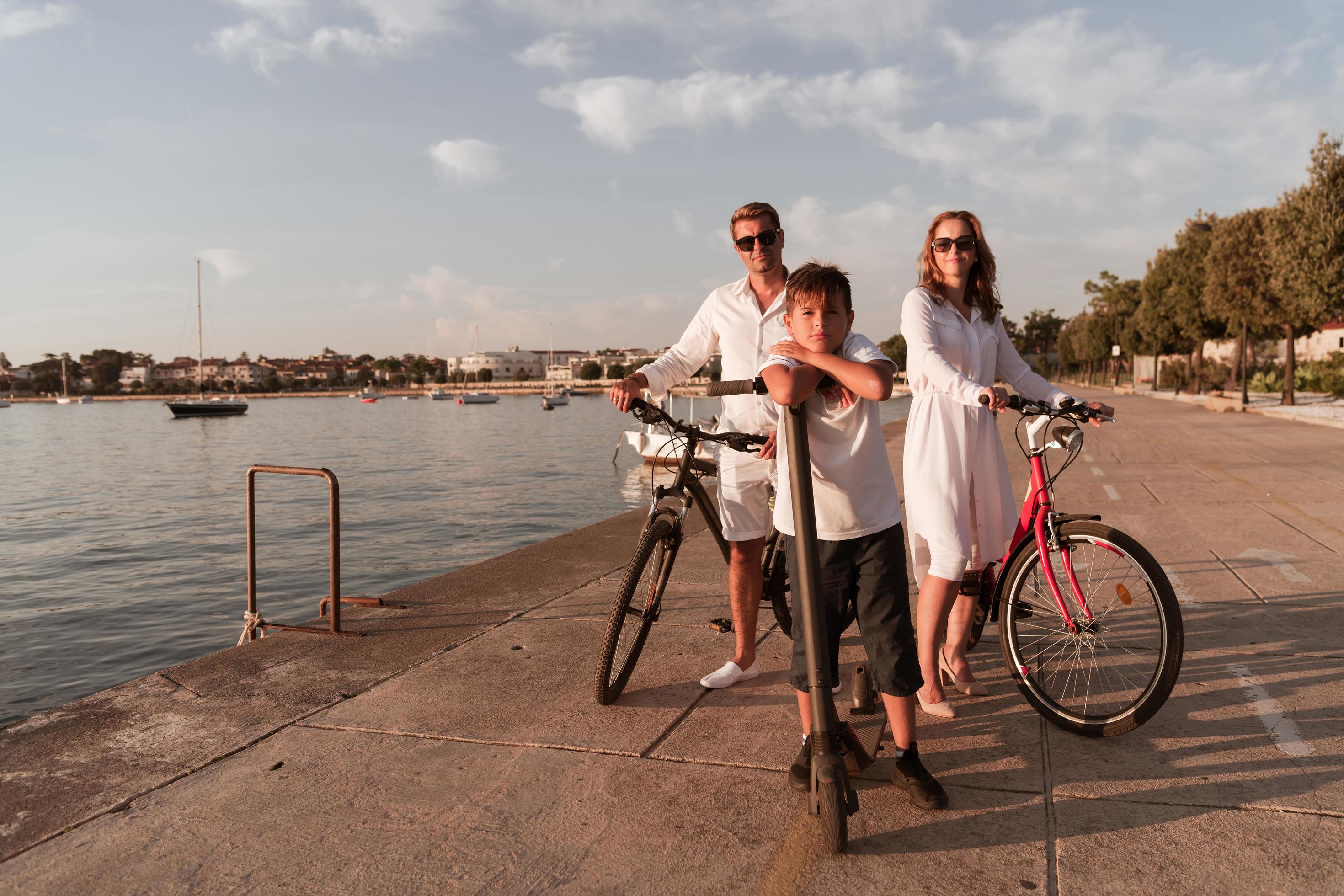 Happy family enjoying a beautiful morning by the sea together, parents riding a bike and their son riding an electric scooter. Selective focus Stock Free