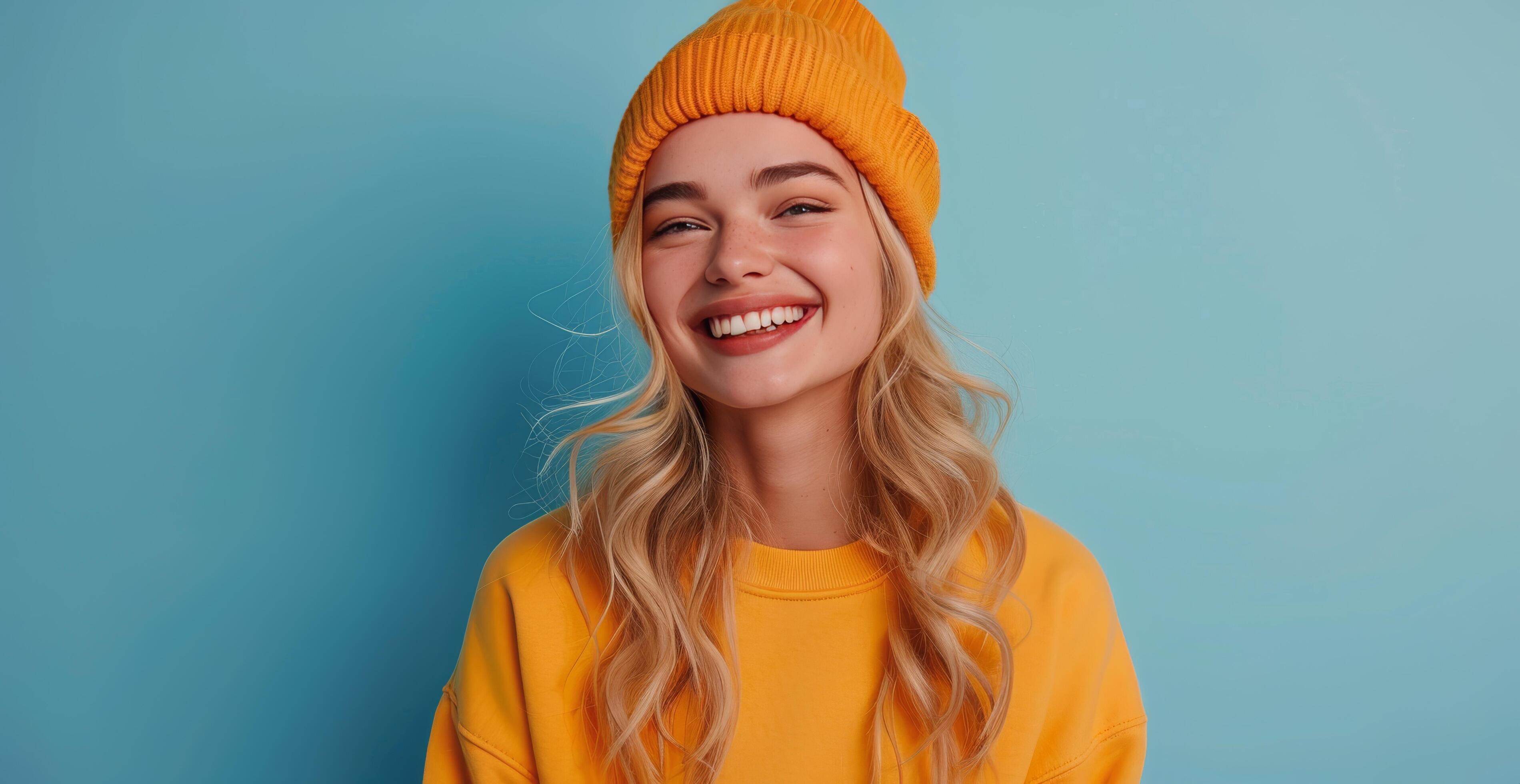 Smiling Young Woman Wearing a Yellow Hoodie and Beanie Against a Blue Background Stock Free